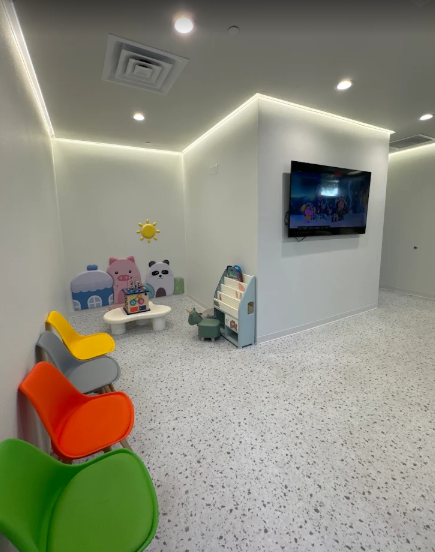 Children's play area with colorful chairs, small tables, and plush animal toys on the wall. A flat-screen TV is mounted on a partition wall, and the room has white walls with recessed lighting.