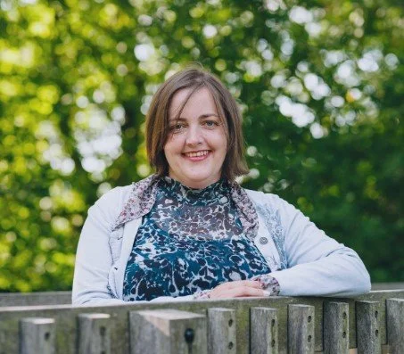 Woman with shoulder-length brown hair smiling, wearing a patterned top and white cardigan, leaning on a wooden fence outdoors with green trees in the background.