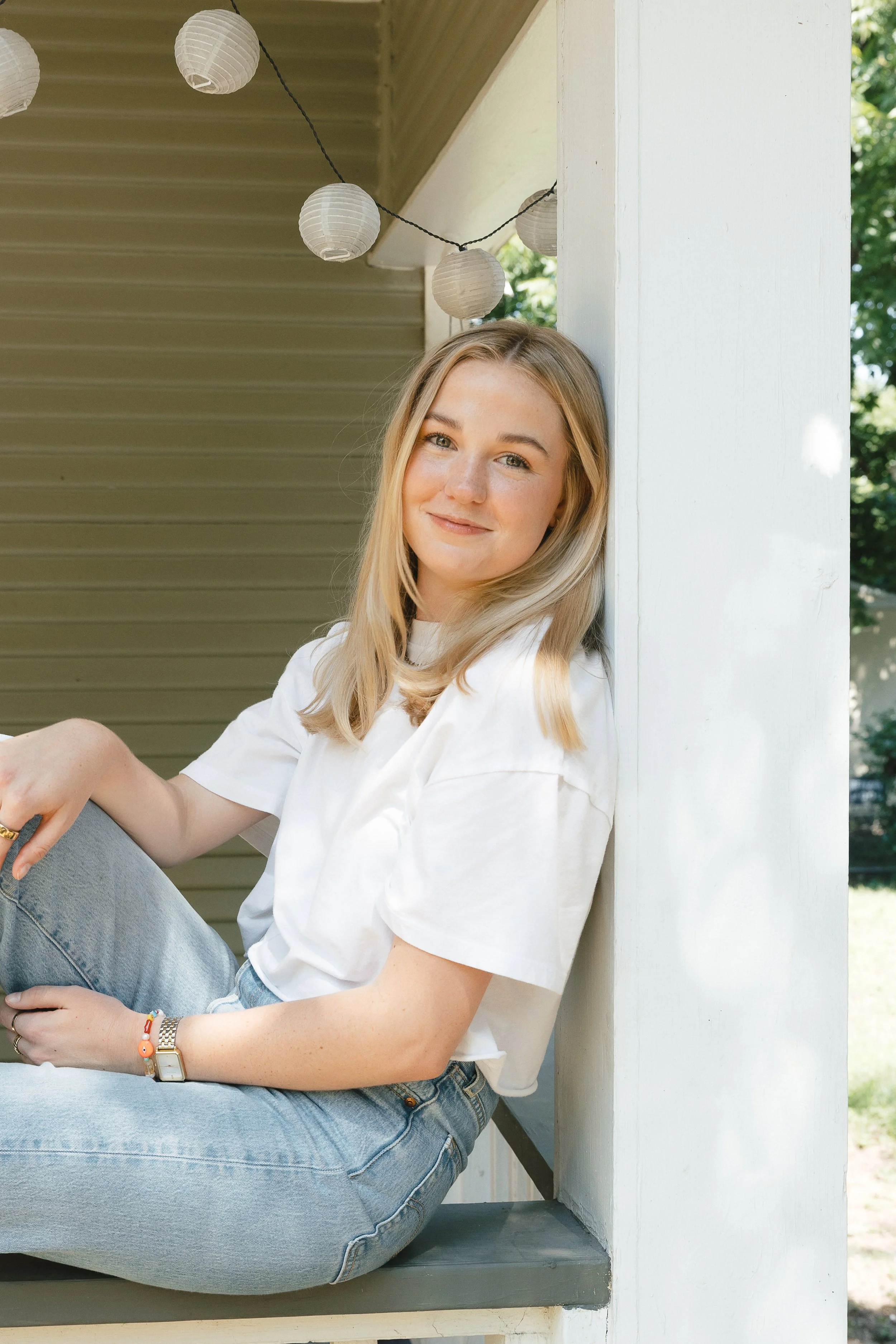 A young woman with long blonde hair smiling while sitting on a beige sofa near a window with cream-colored curtains.