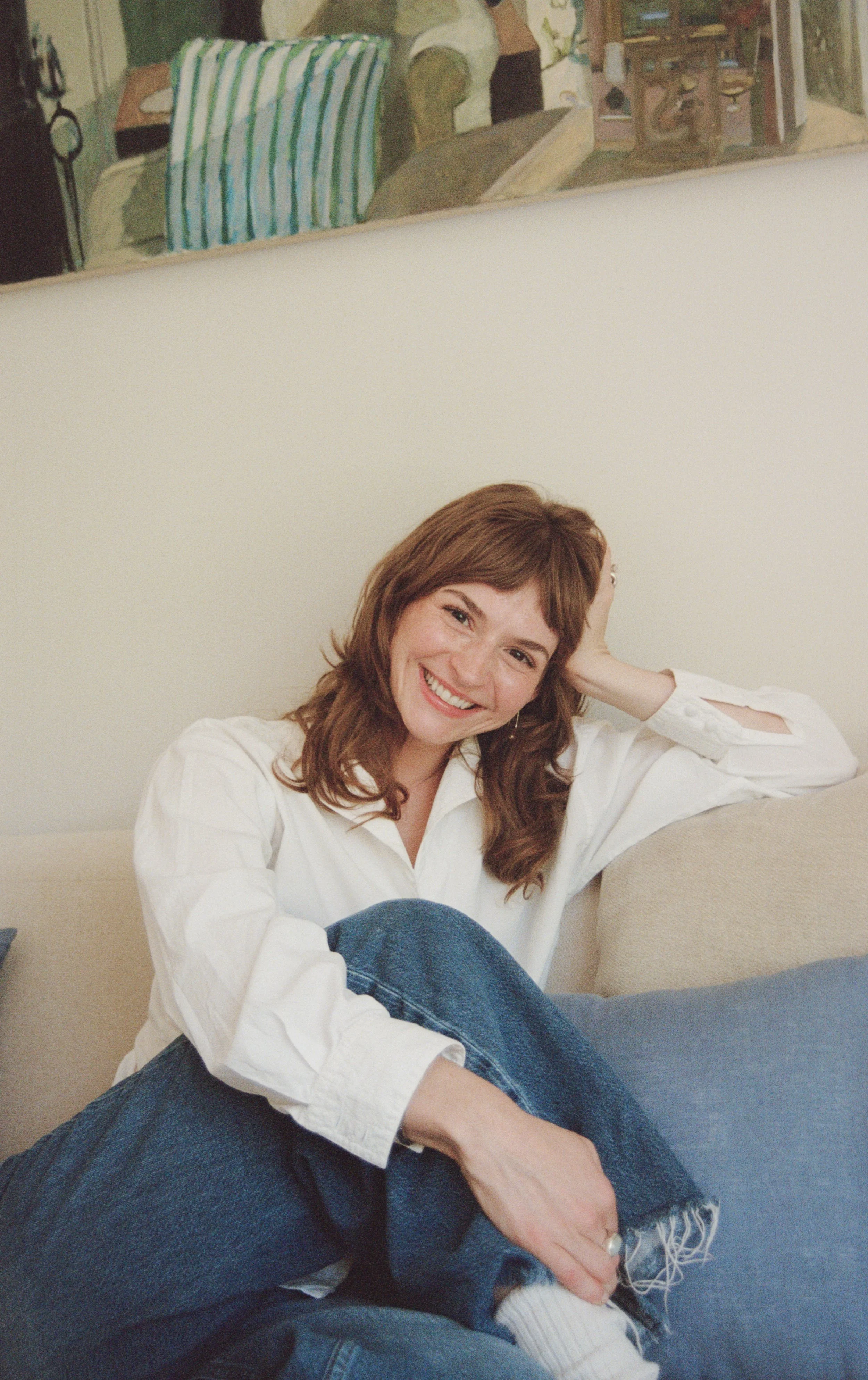 A woman with short light brown hair and blue eyes resting her head on a cream-colored sofa cushion near a window with sheer curtains, smiling softly.