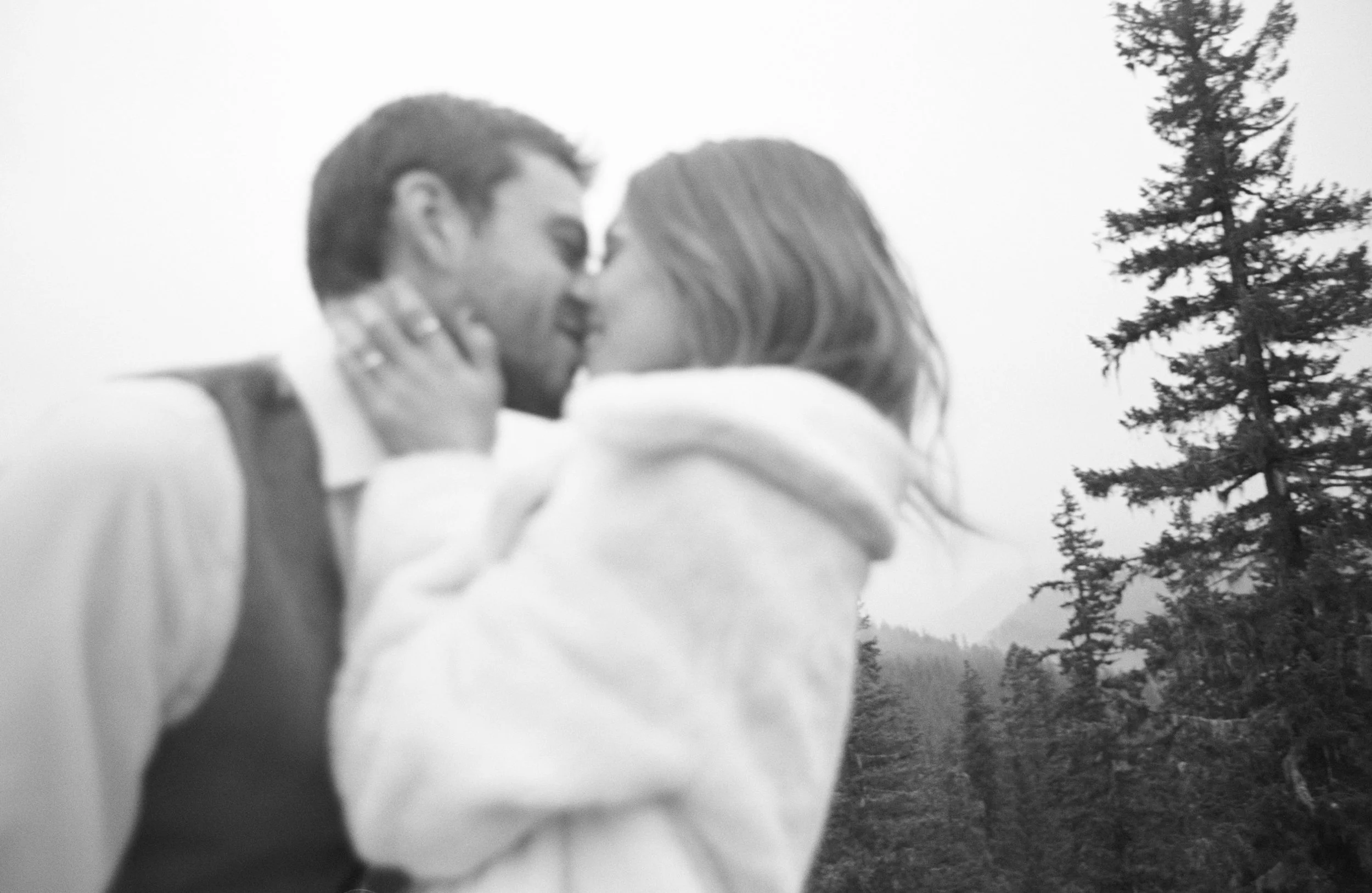 A black-and-white photograph of a couple kissing outdoors with a forest and trees in the background.