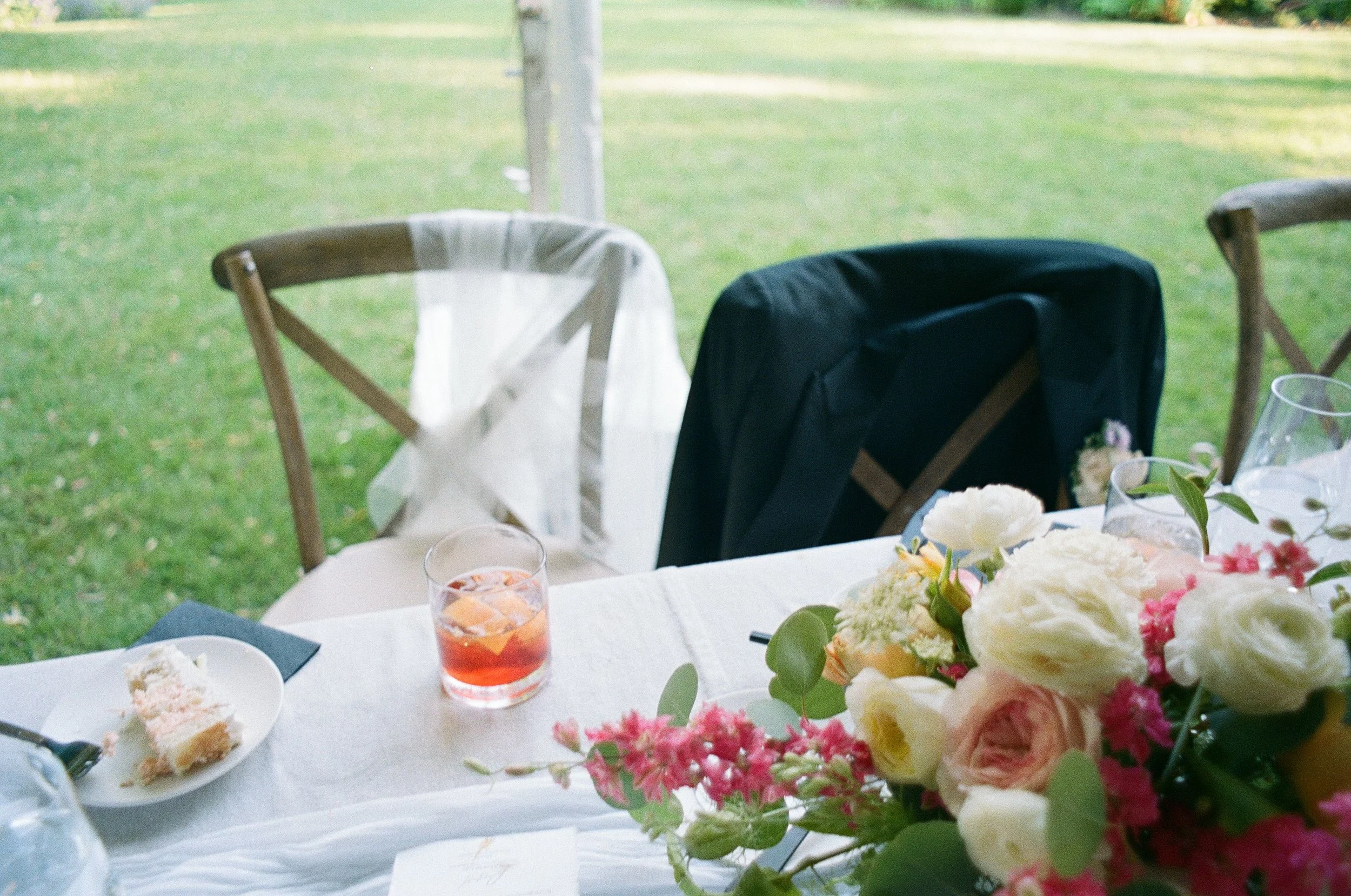 A decorated table set for an outdoor event with a white tablecloth, a floral centerpiece with white and pink flowers, and drinks. There are three chairs around the table, one with a white fabric draped over the back, and another with a black cover. T