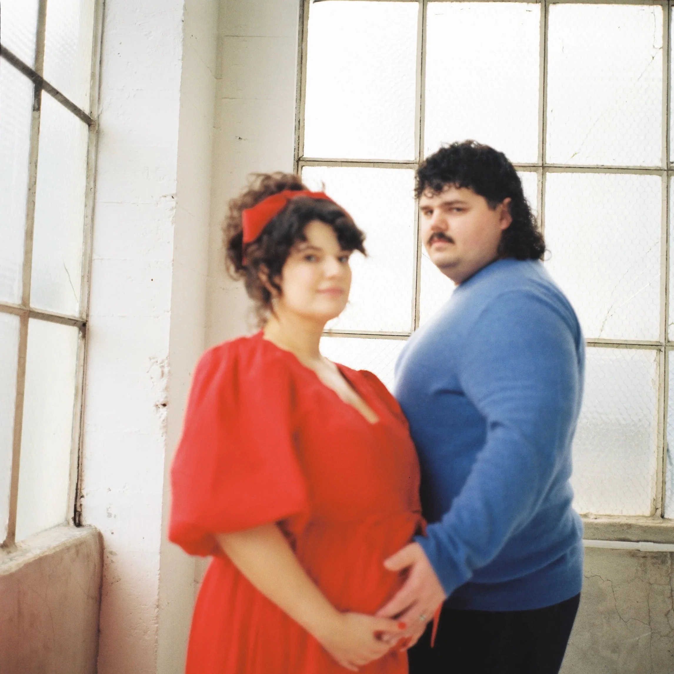 A couple holding their pregnant belly, standing inside a room with large industrial windows in the background. The woman is wearing a red dress and a red headband, the man is wearing a blue sweater and has a mustache.