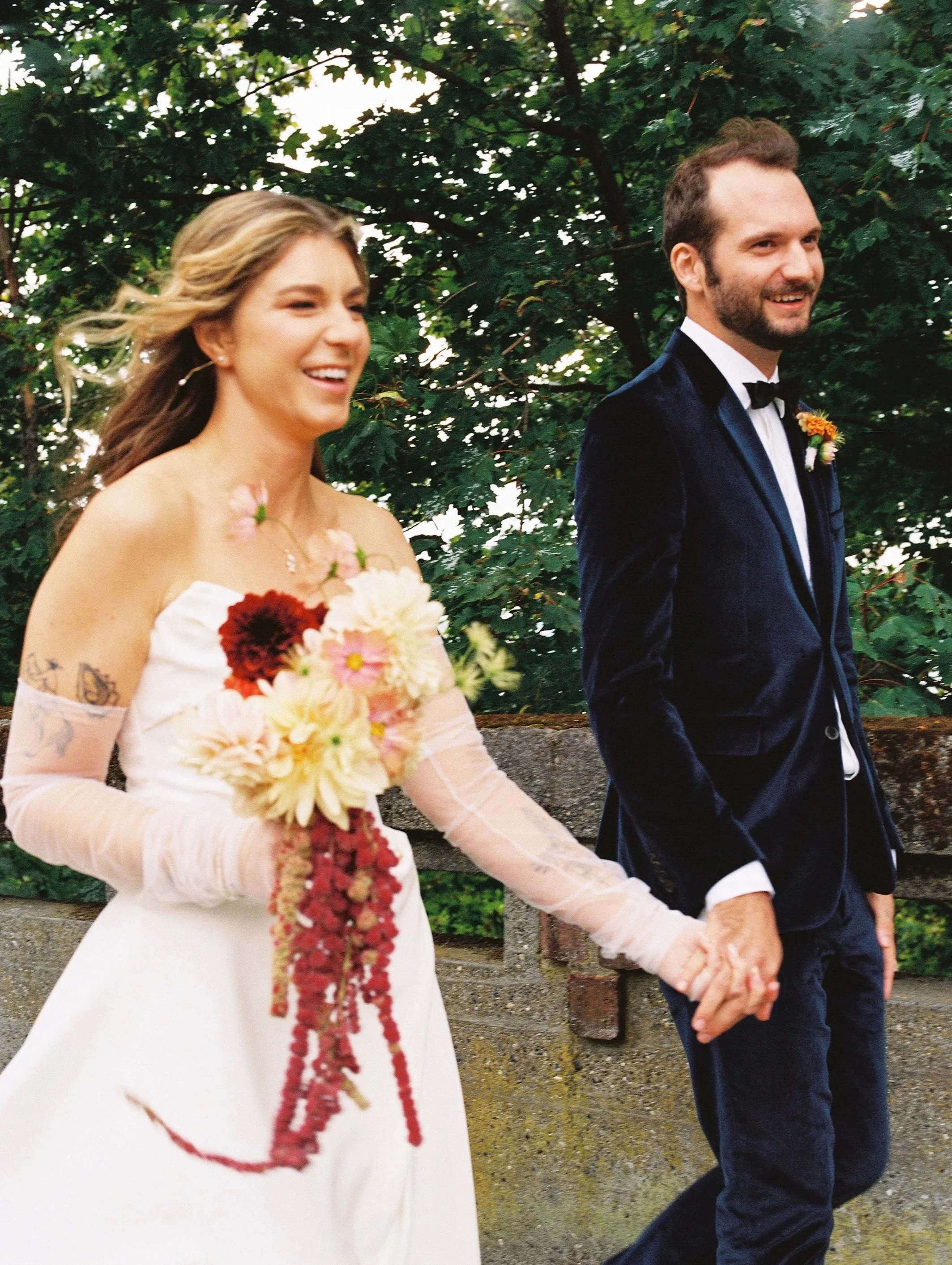 A happy bride and groom holding hands and smiling outdoors on their wedding day, with green trees in the background.