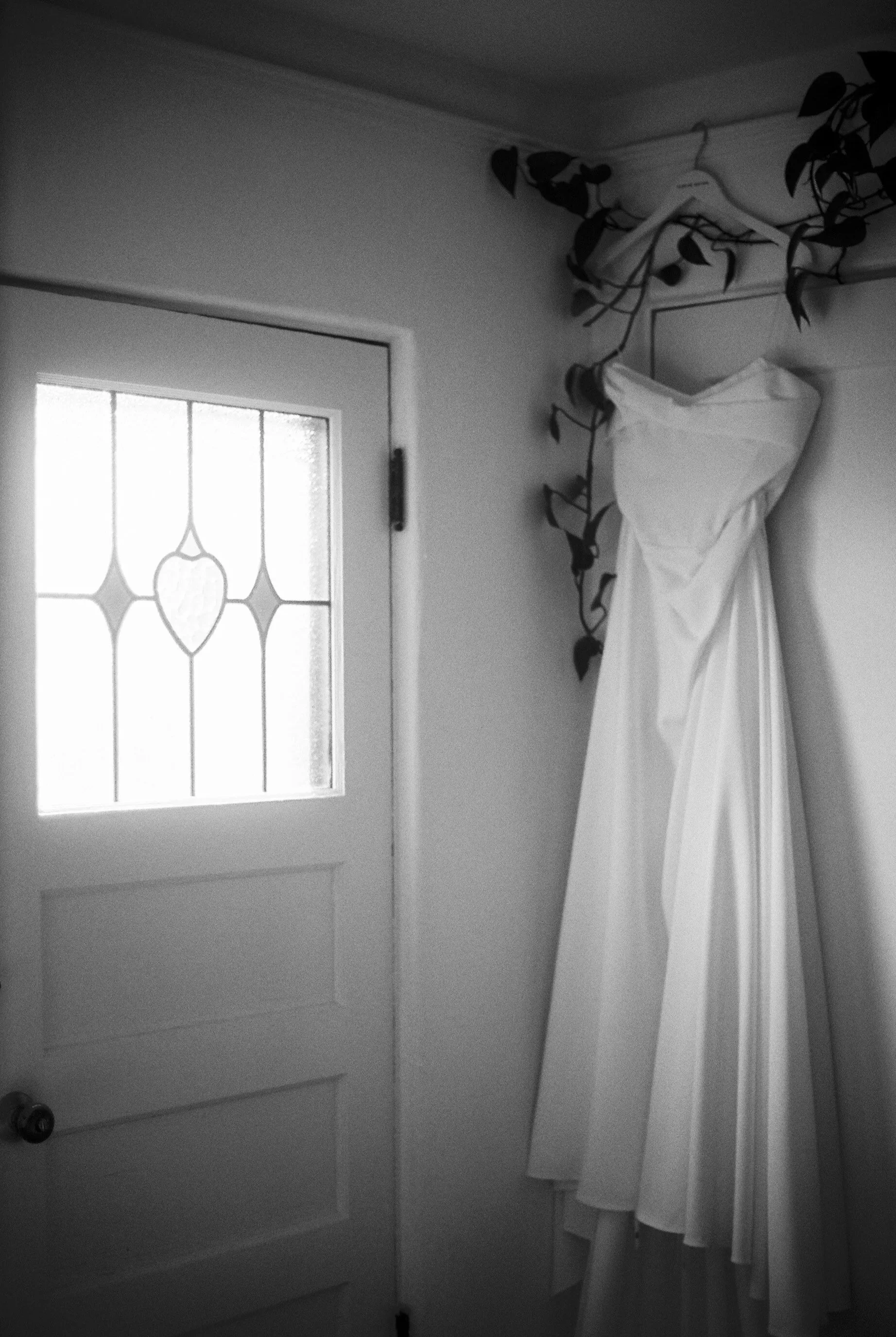 A white dress hanging on a hanger on a wall beside a decorative vine, near a door with a stained glass window.