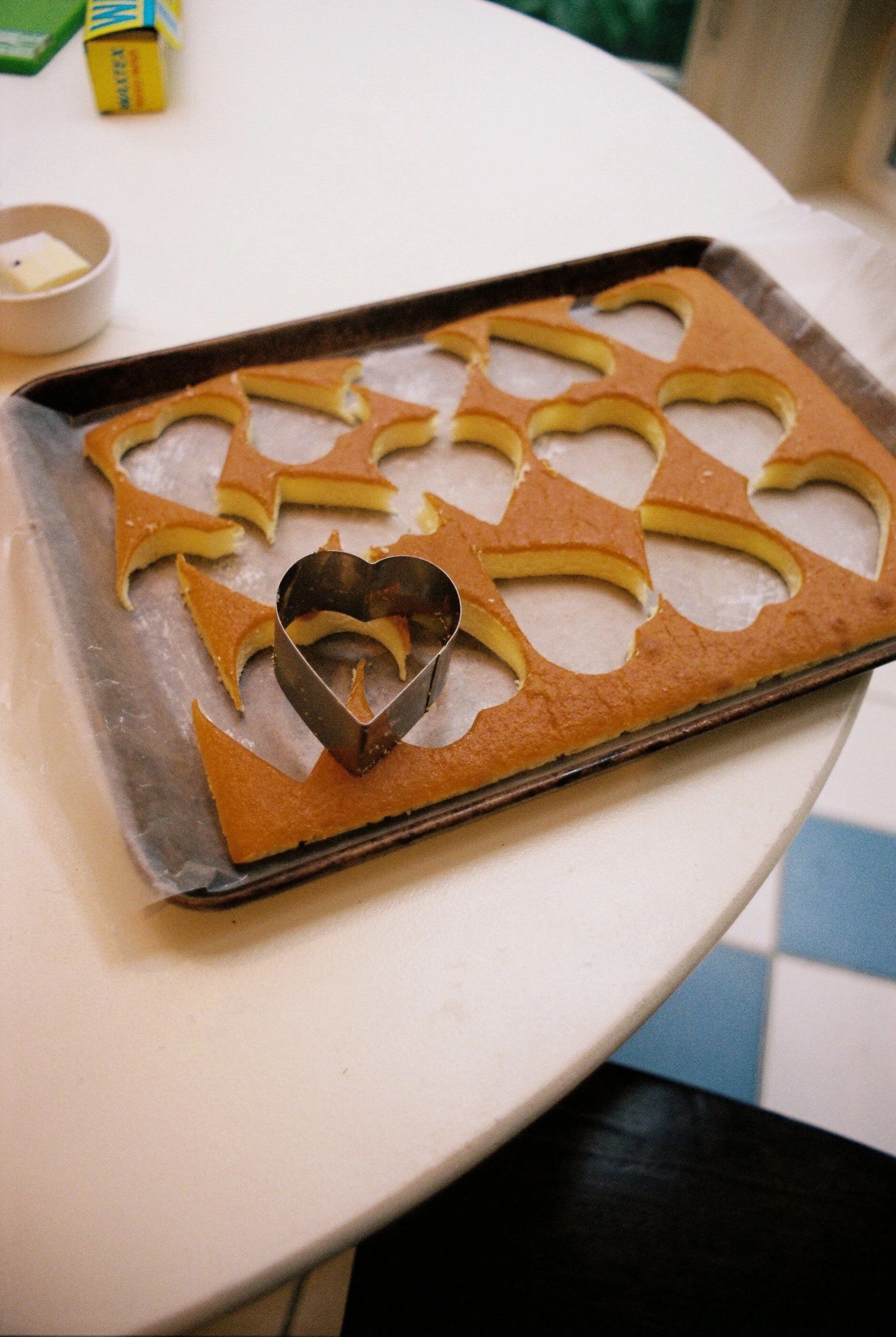 A baking tray with heart-shaped cutouts from a sheet of cake, and a heart-shaped metal cookie cutter placed on top, sitting on a white table.