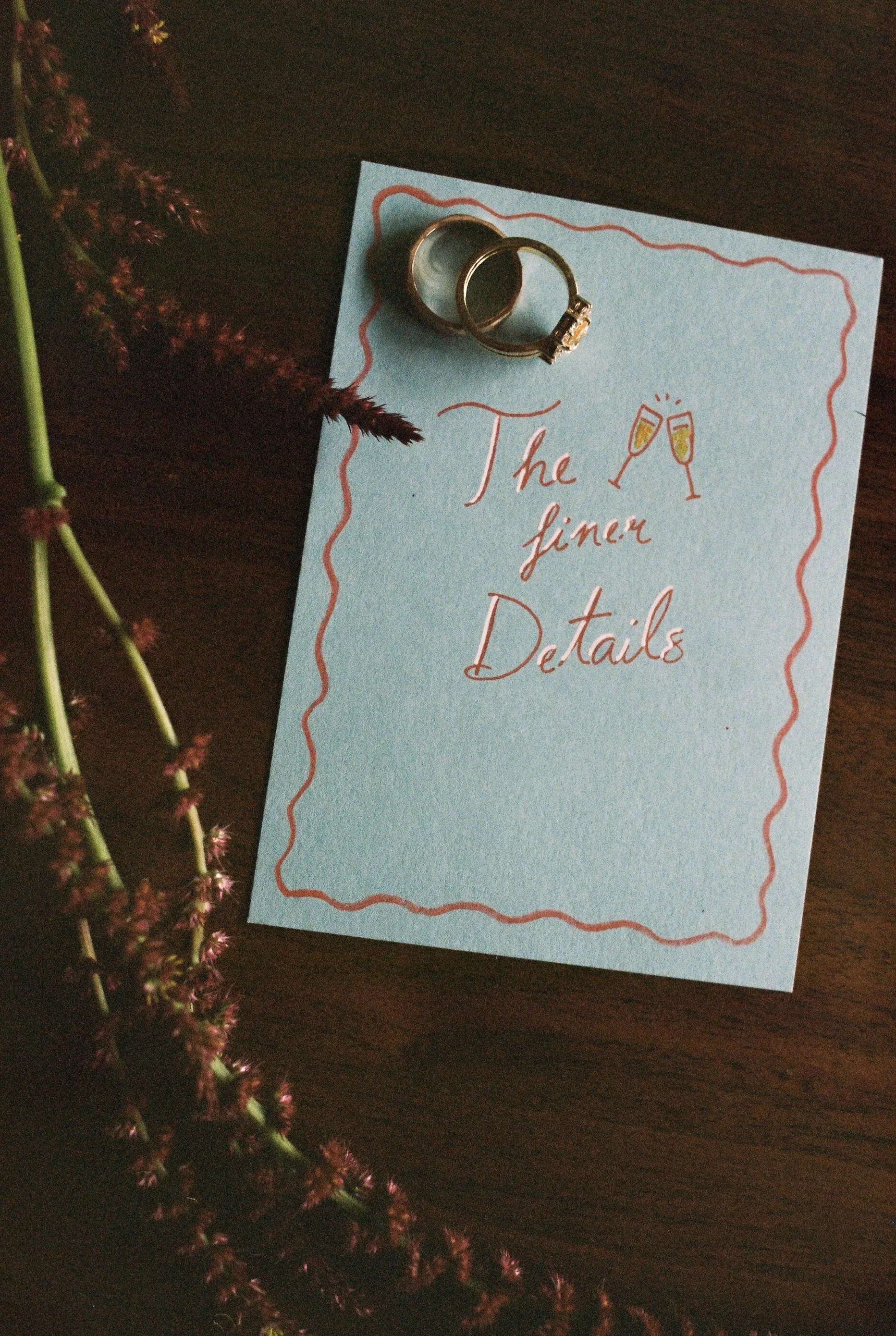 A light blue card with red borders and handwritten text that reads 'The finer Details' with a drawing of two champagne glasses clinking. Two rings, one with a large stone, are placed on the card. A sprig of purple flowers is partially visible on the 