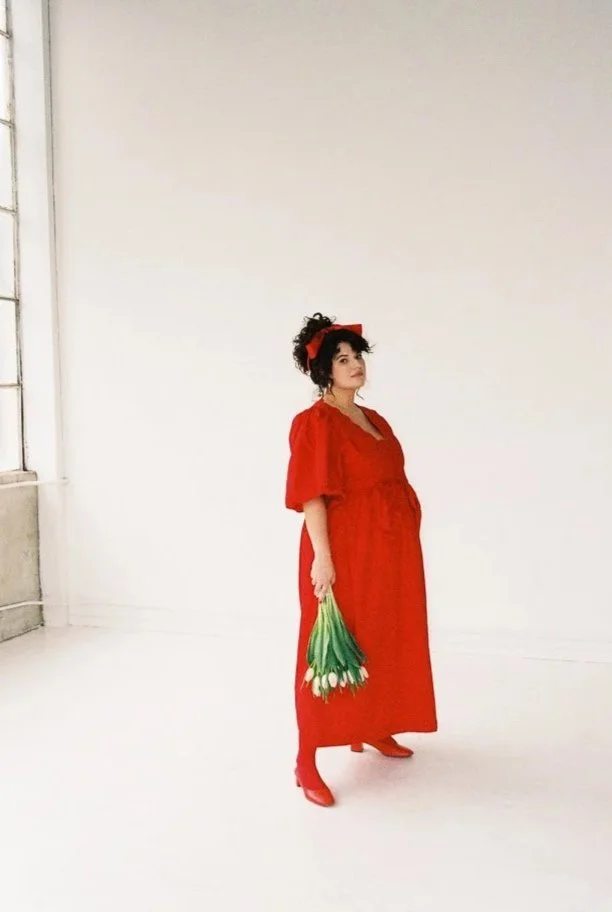 A woman wearing a red dress and high heels, holding a bouquet of white tulips, standing in a minimalistic room with white walls and floor.