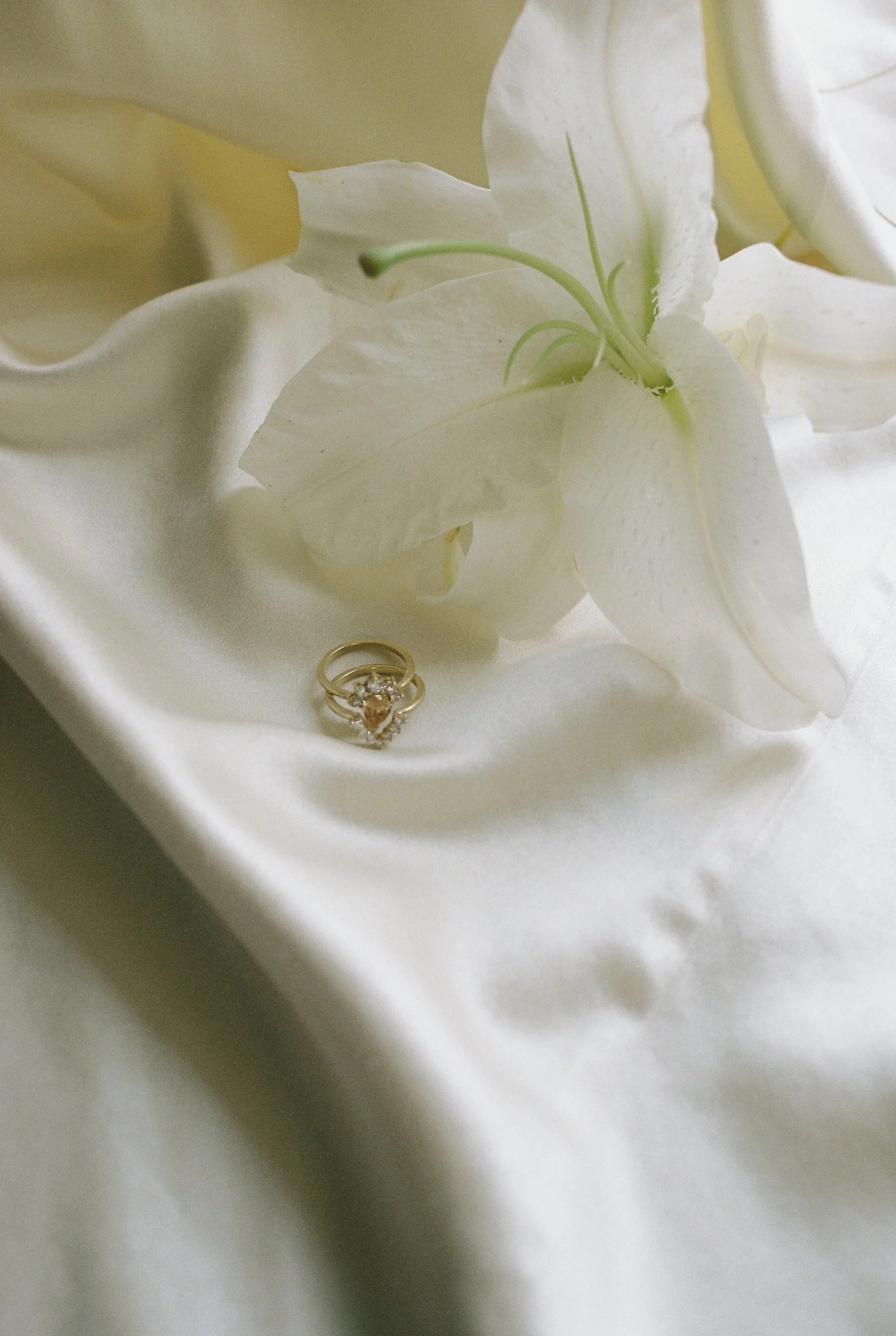 A close-up of a white lily flower with two gold rings, one with a heart-shaped gemstone, resting on white satin fabric.