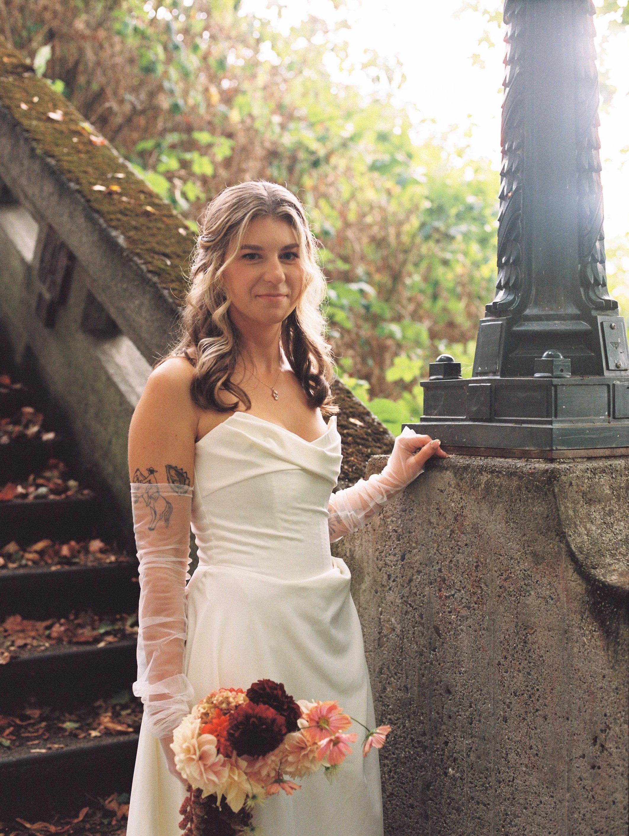 A bride in a white wedding dress holding a bouquet of flowers, standing outdoors near a stone railing, with lush green trees and sunlight in the background.