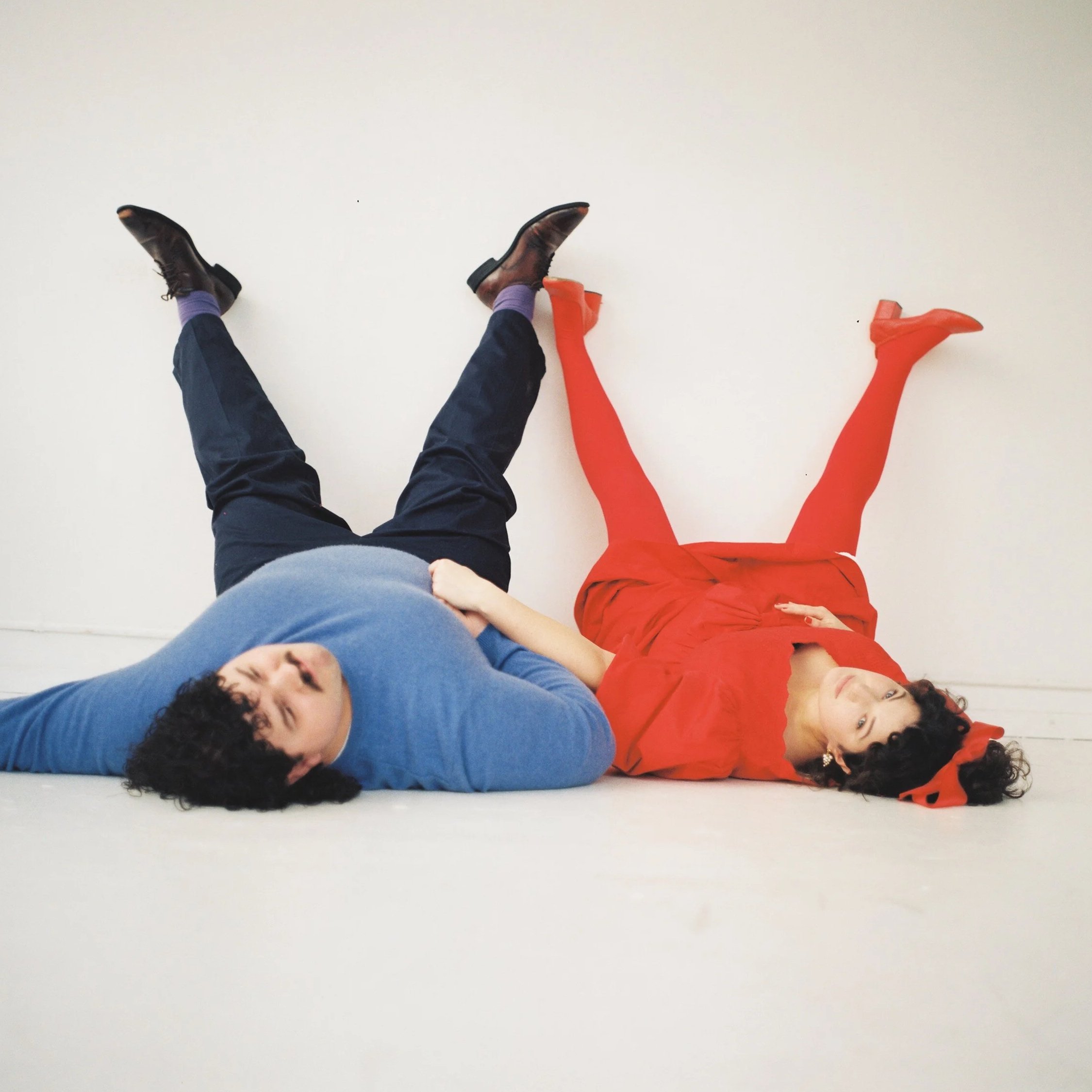 Two women lying on a white floor with their legs raised against a white wall, one in a blue long-sleeve shirt and the other in a red dress with matching red tights.