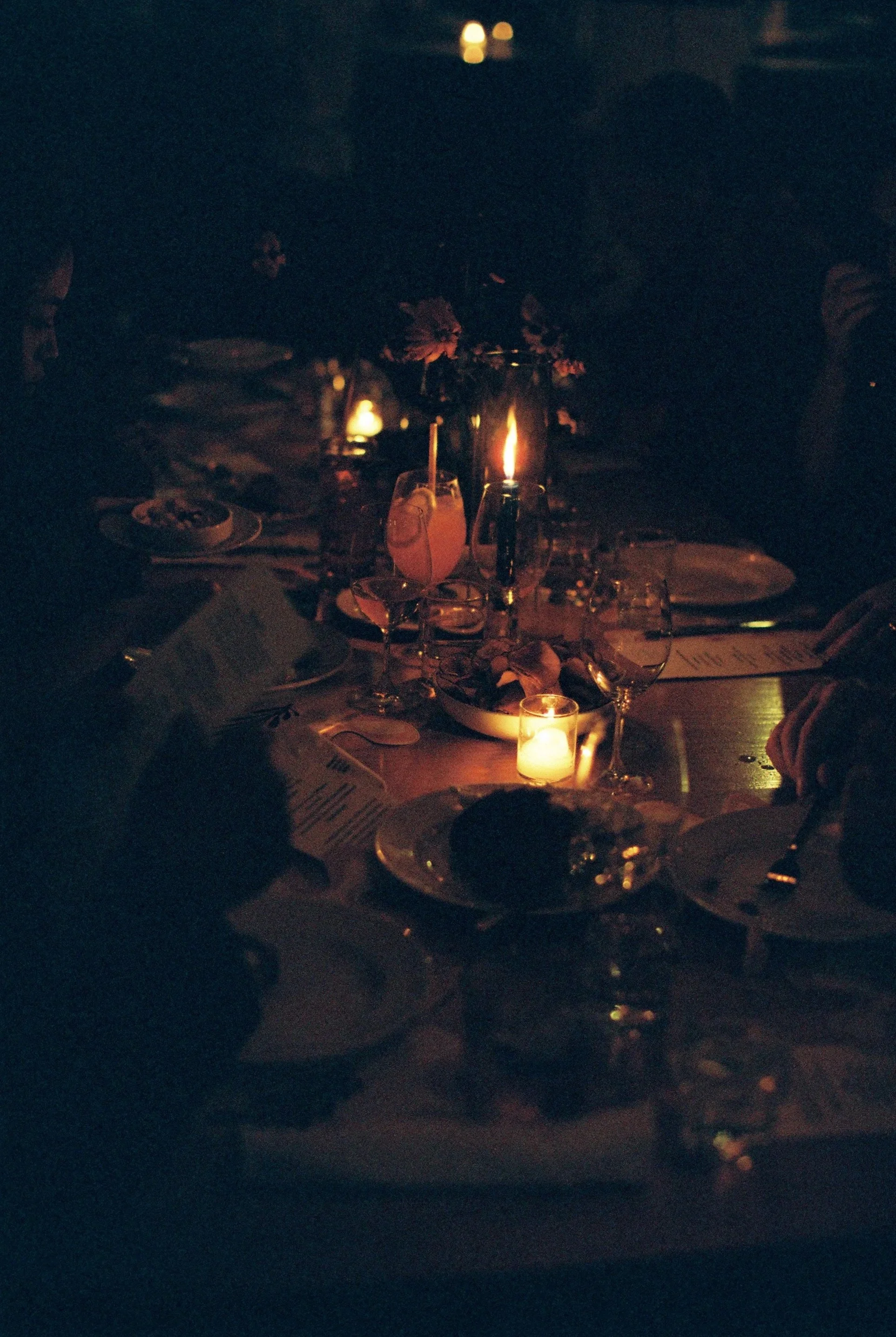 A darkly lit dining table with candles, floral centerpieces, glasses, plates, and menus, set for a meal in an intimate setting.