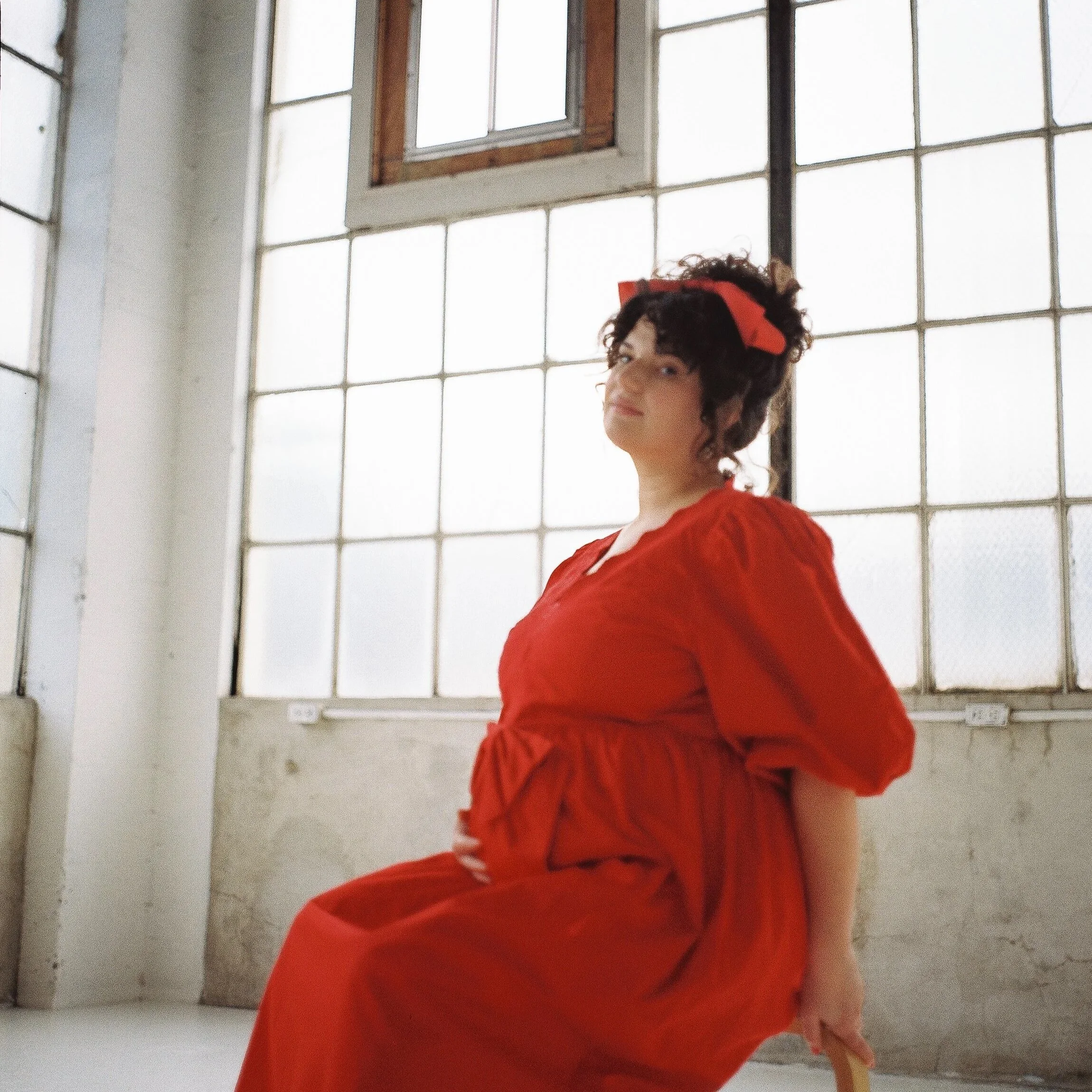 A woman with curly hair wearing a red dress and red headband sitting on a wooden chair in front of large industrial windows in a bright, minimalist room.