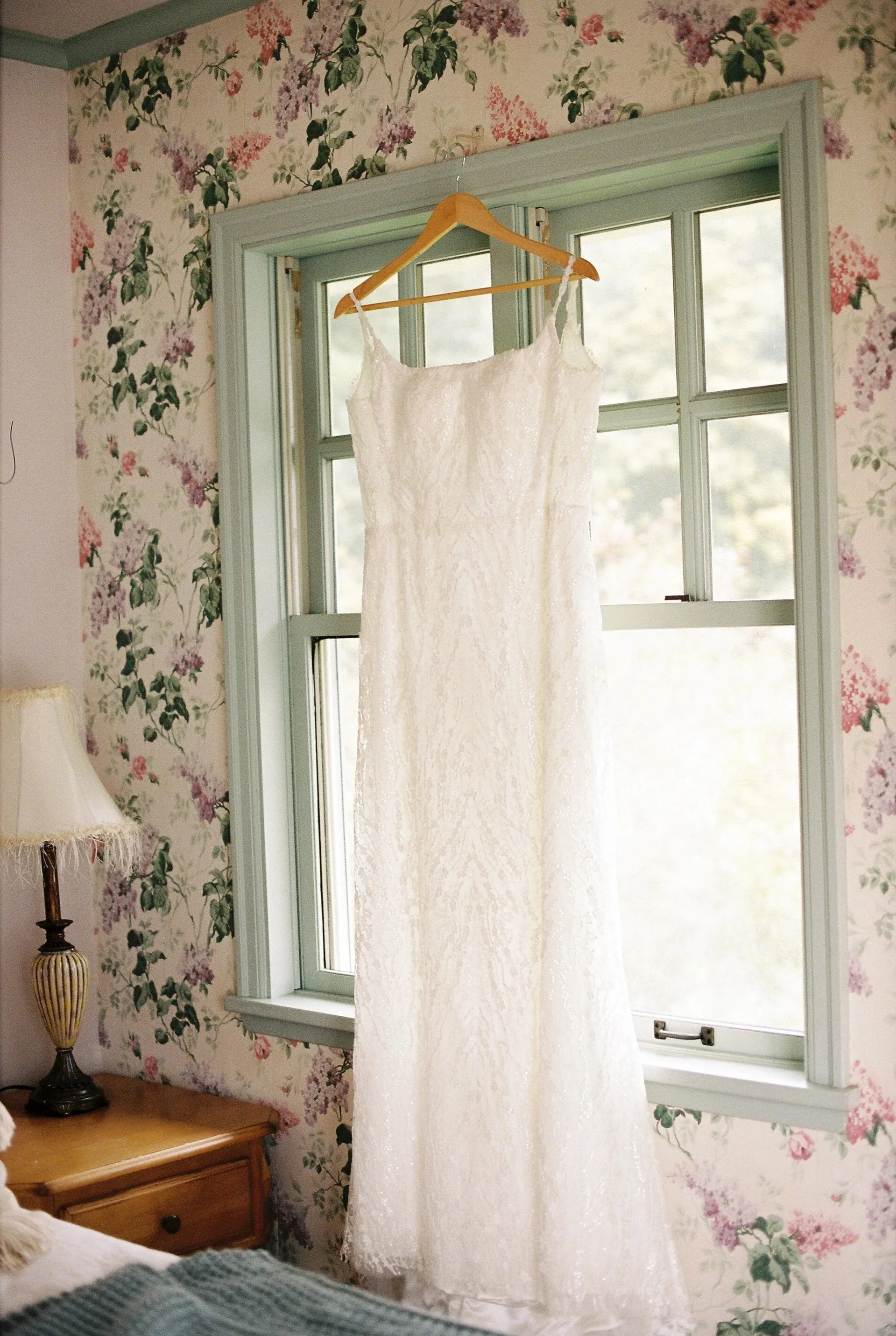 A white wedding dress hanging from a hanger on a window frame in a bedroom with floral wallpaper.