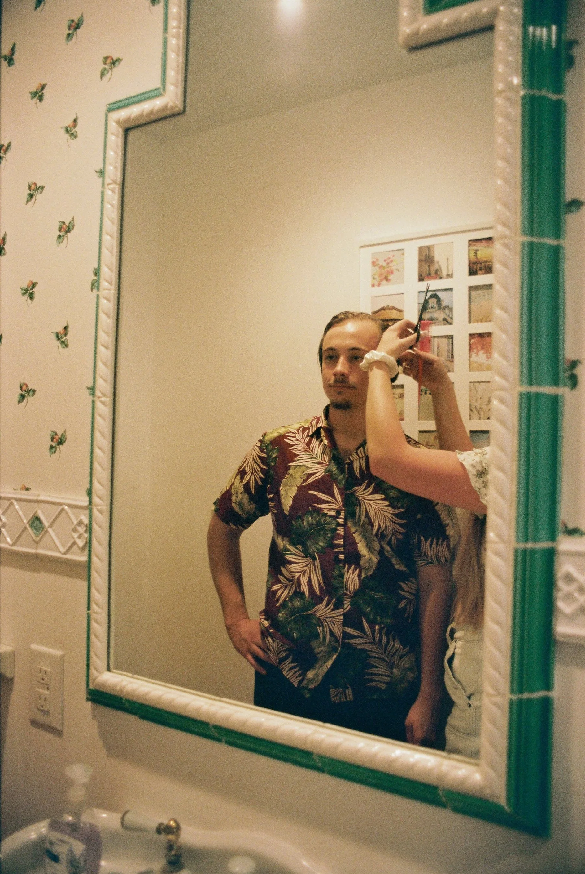 A man stands in front of a bathroom mirror with a person behind him trimming his hair. The man has a mustache and wears a colorful Hawaiian shirt. The bathroom wall has a floral wallpaper pattern, and there are framed pictures on the wall behind him.