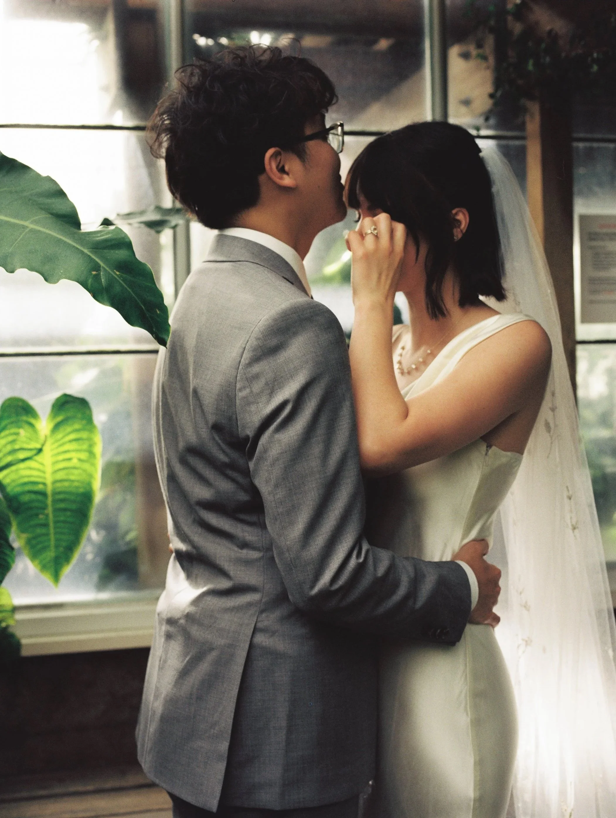 A couple in wedding attire, sharing an intimate moment indoors near a window with greenery outside.