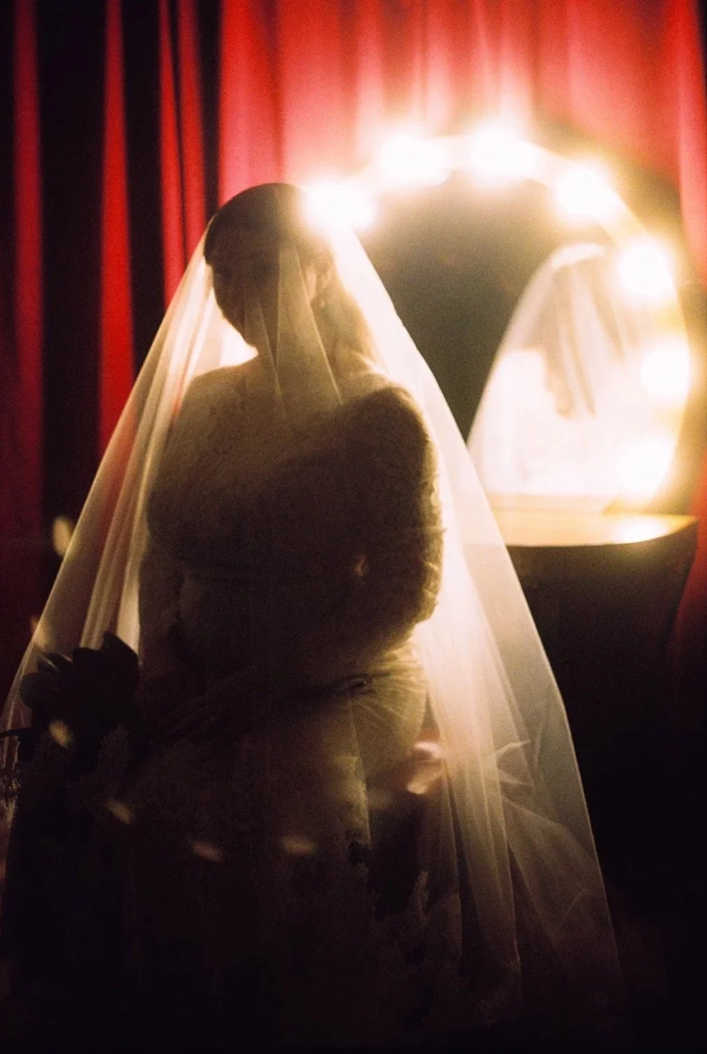 A bride in a wedding dress and veil standing in front of a brightly lit vanity mirror, with red curtains in the background.
