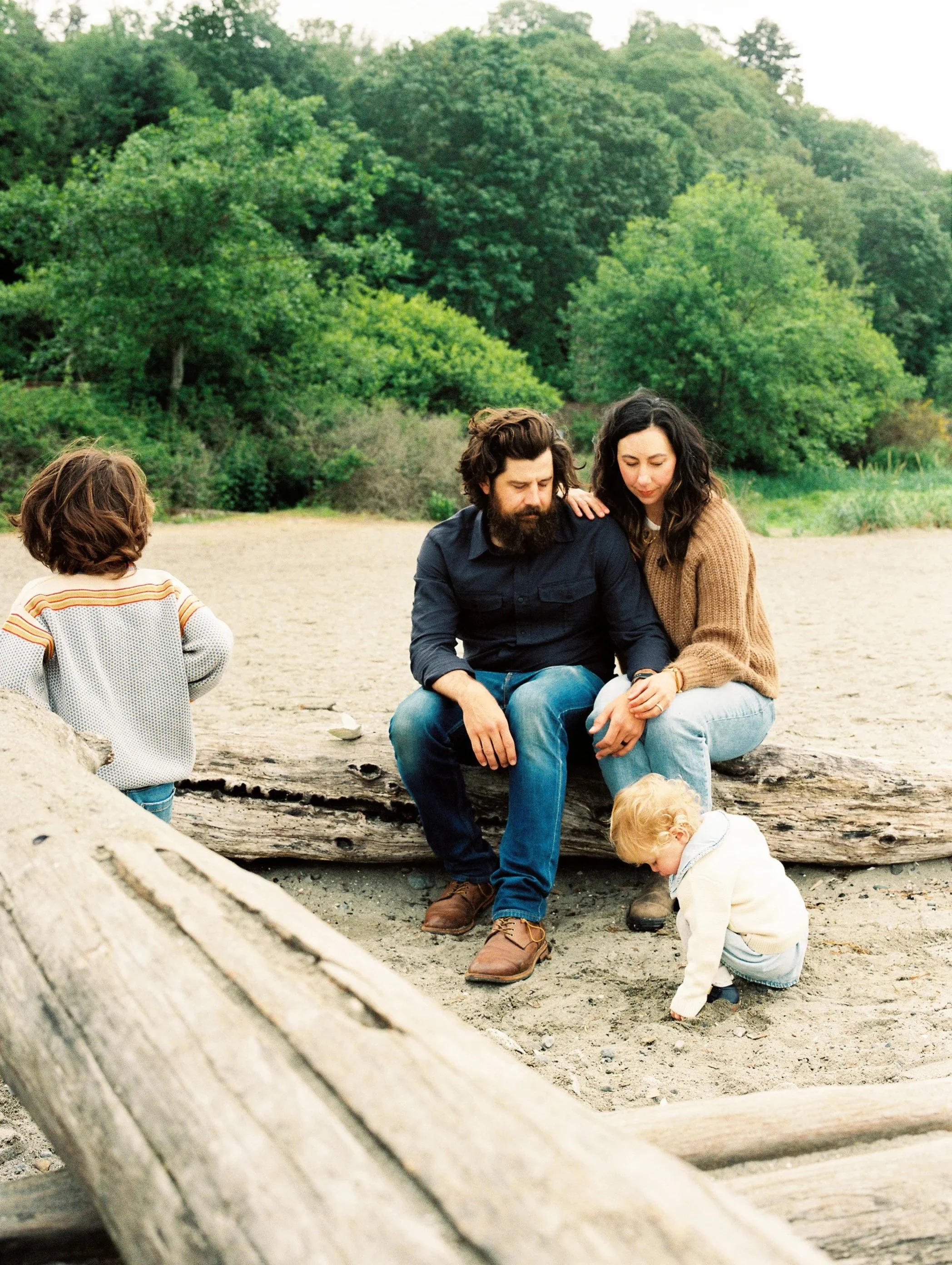 A family of four sitting on a large log on a beach with greenery in the background. The parents are sitting close together, with a young girl standing near the log and a young boy playing in the sand.