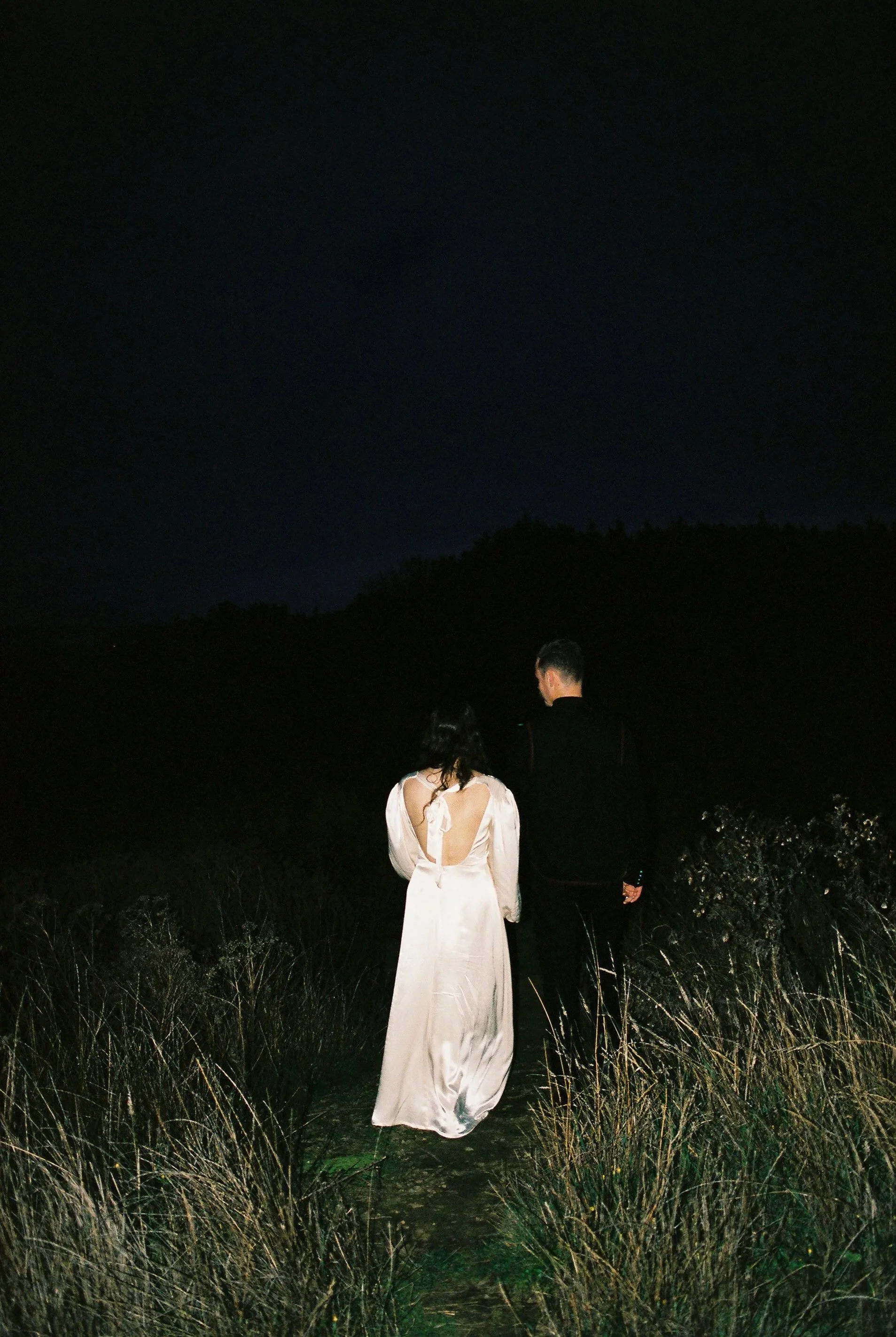 A woman in a long, white satin dress and a man in a black suit walking outdoors at night on a narrow path surrounded by grass.