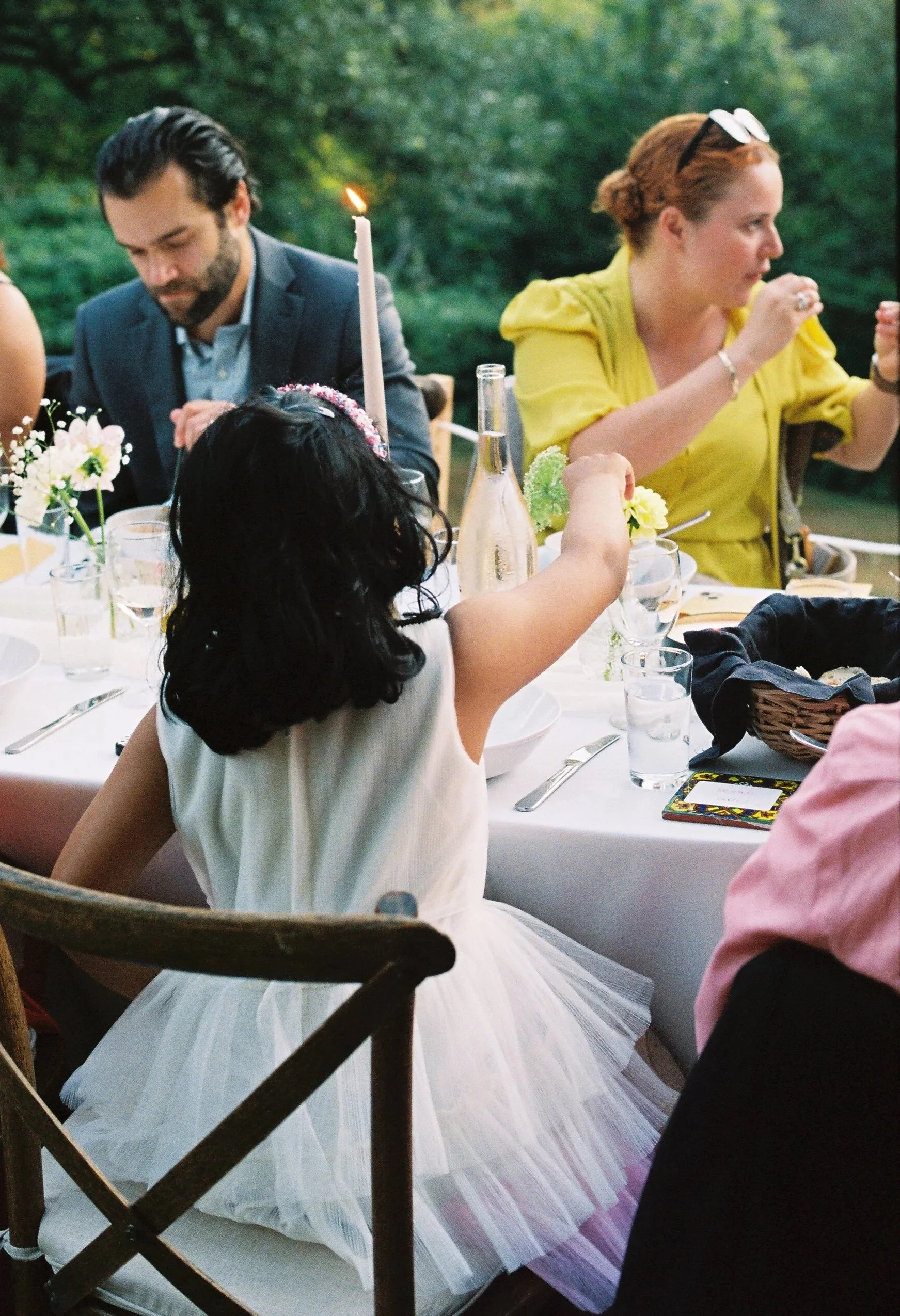 Children and adults sitting at a table outdoors during a gathering or celebration, with decorated food and drinks, and a candle lit in the center.