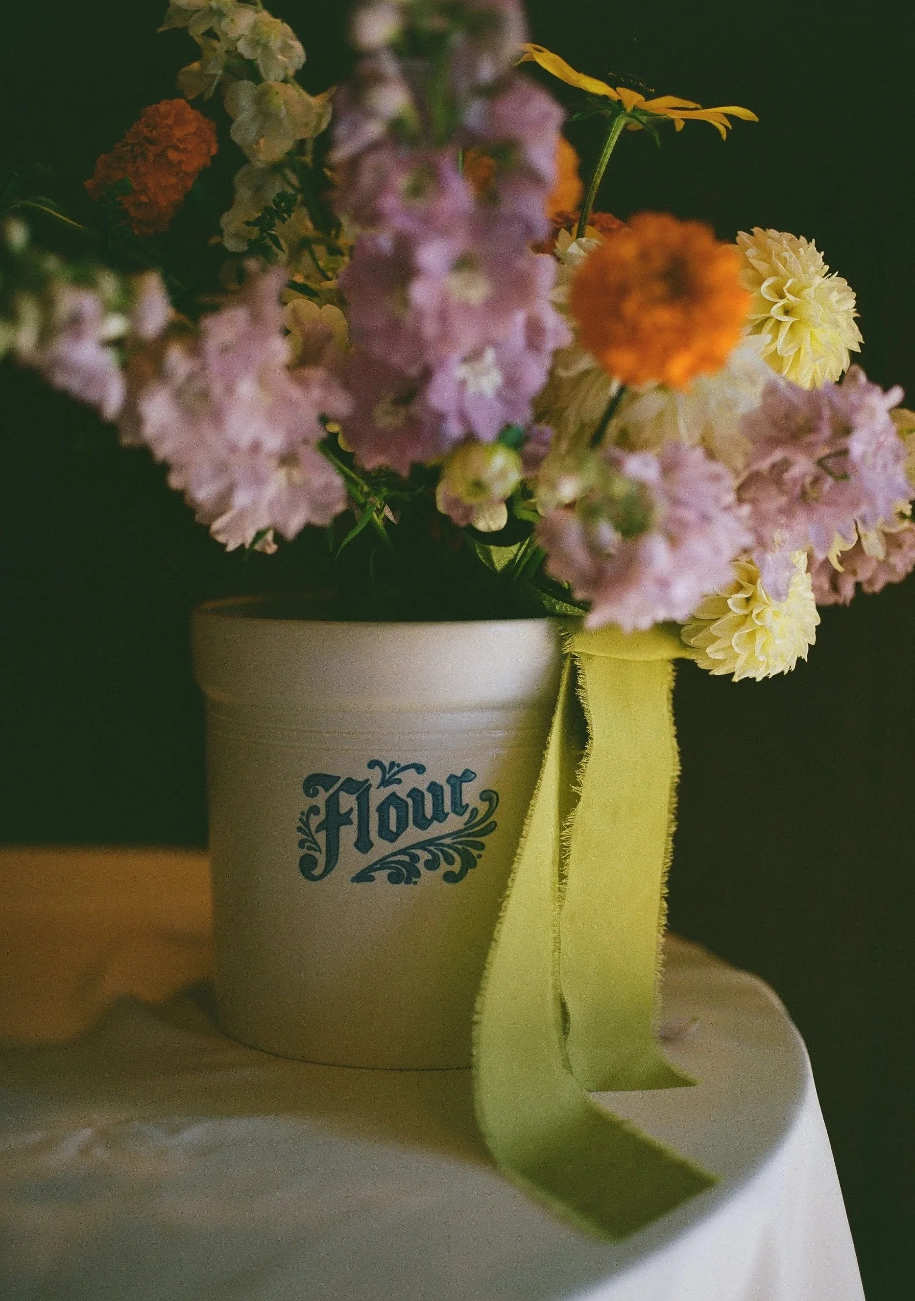 A bouquet of pink, orange, yellow, and purple flowers in a white container labeled 'Flour,' placed on a white table with a yellow ribbon hanging from the container.