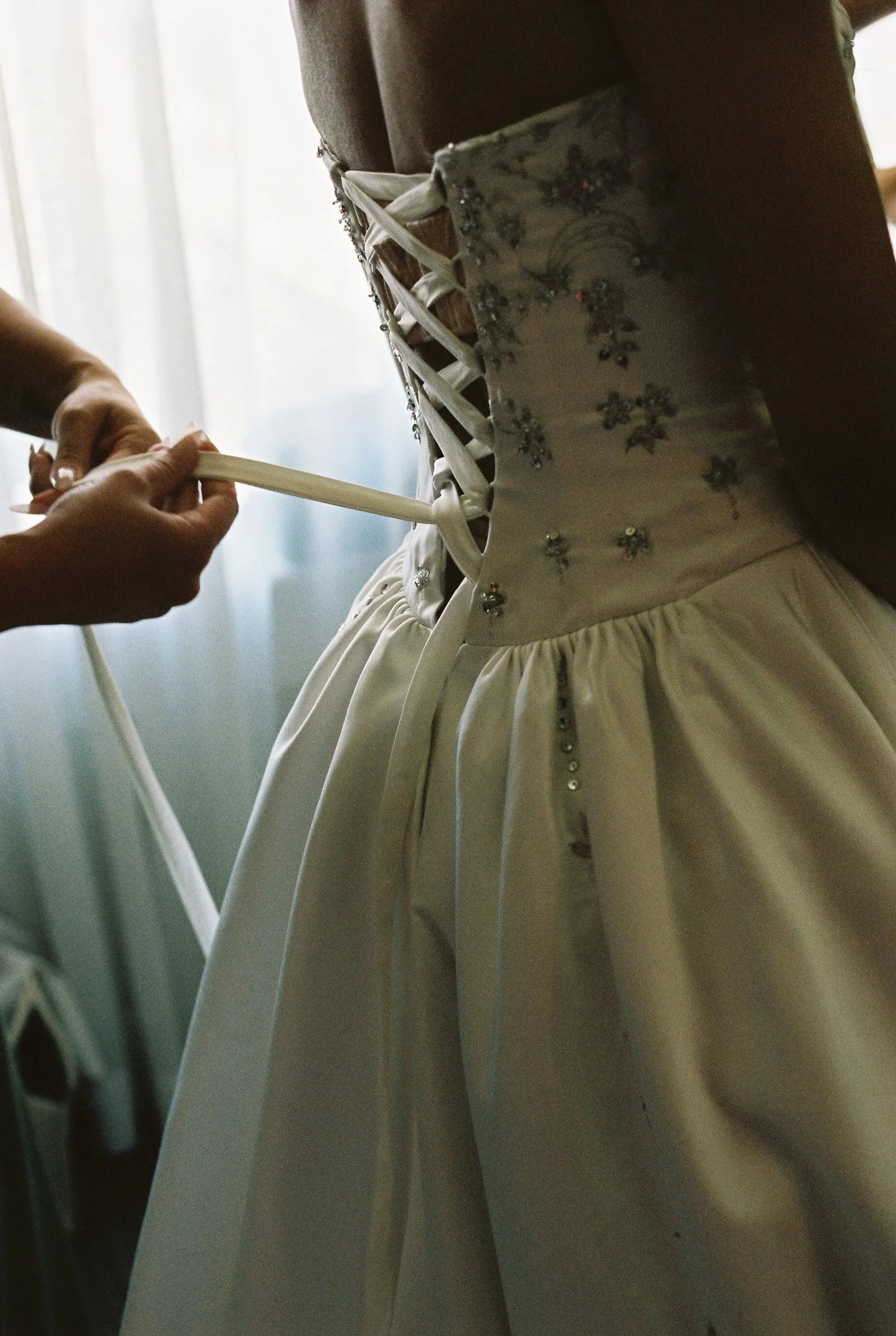 Close-up of someone lacing up the back of a white wedding dress with floral embroidery and beadwork.
