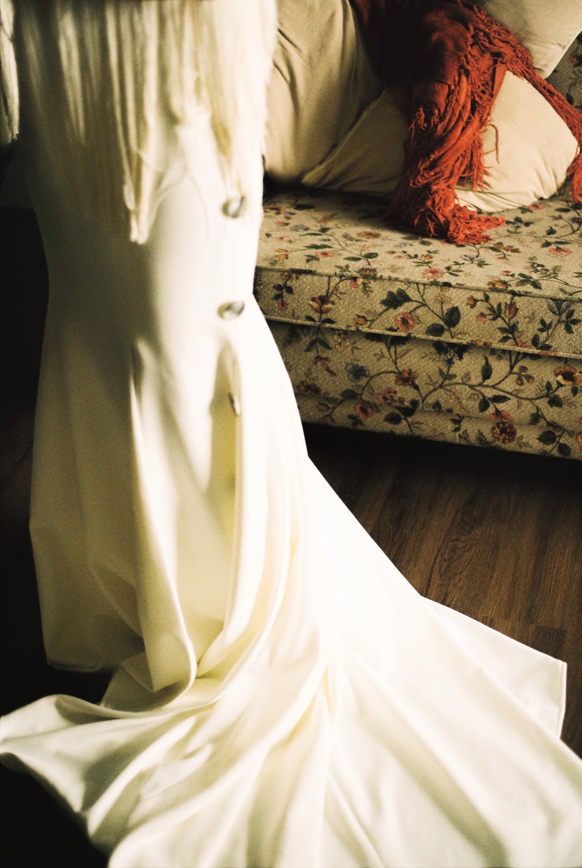 A close-up of a white wedding dress with buttons down the back, draped on a surface next to a floral patterned sofa with pillows and a red scarf.
