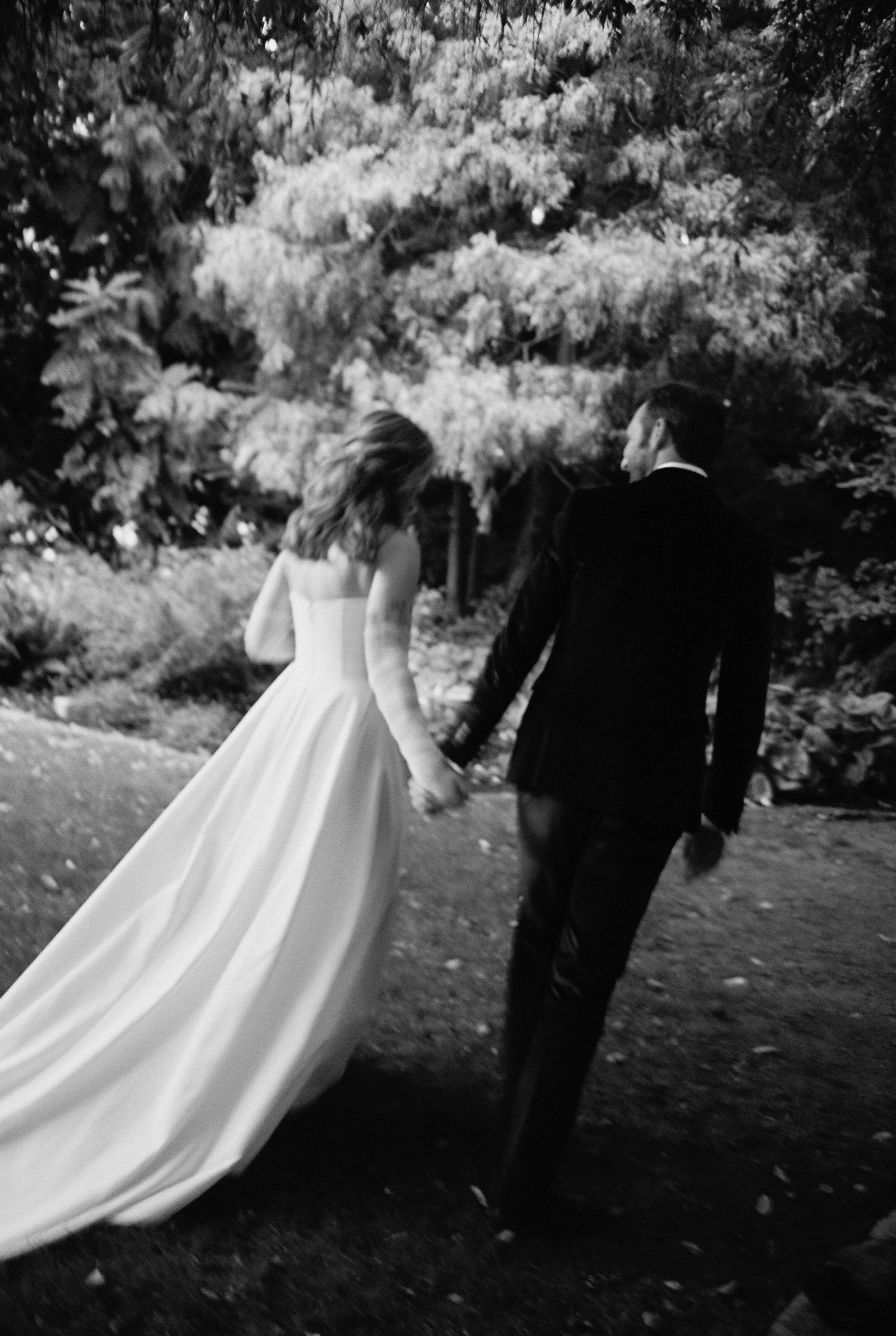 A black-and-white photo of a bride and groom holding hands and walking outdoors, with trees and foliage in the background.