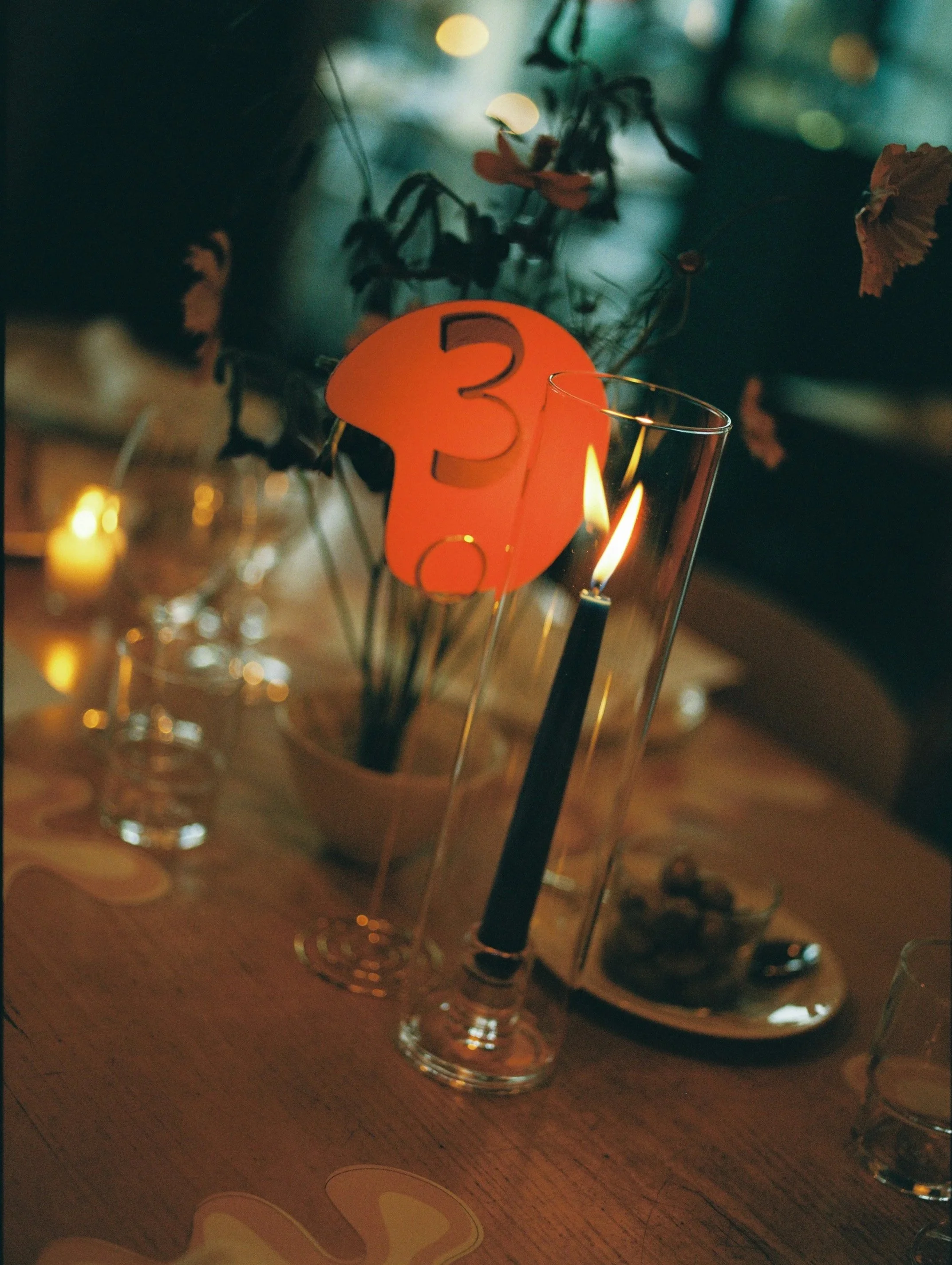 A lit black candle in a tall glass holder, orange table number sign with the number 3, and a floral centerpiece with pink flowers on a wooden table at a dimly lit celebration or event.
