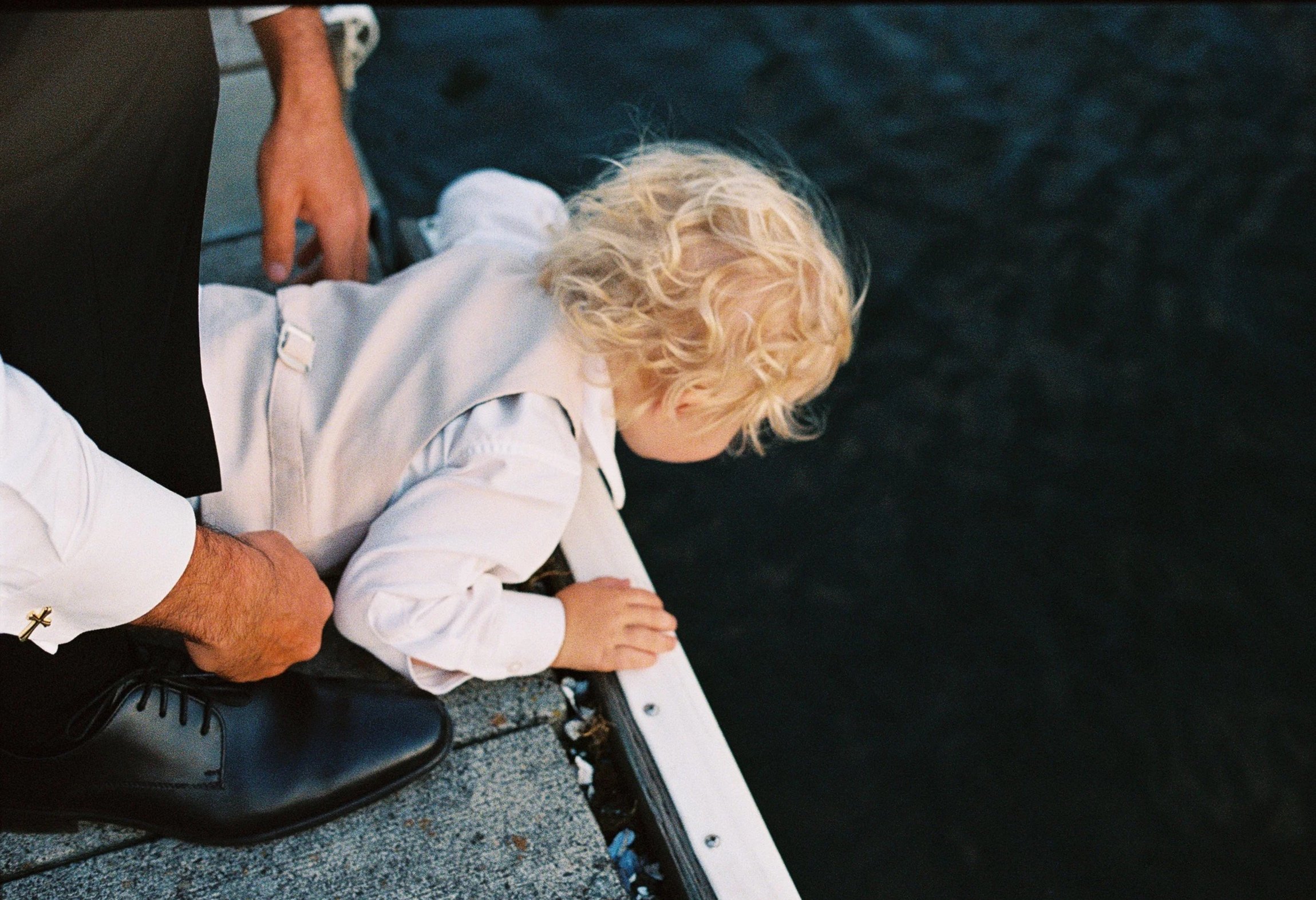 A young child with curly blonde hair leans over the edge of a pier, looking into the water below, with an adult standing nearby.