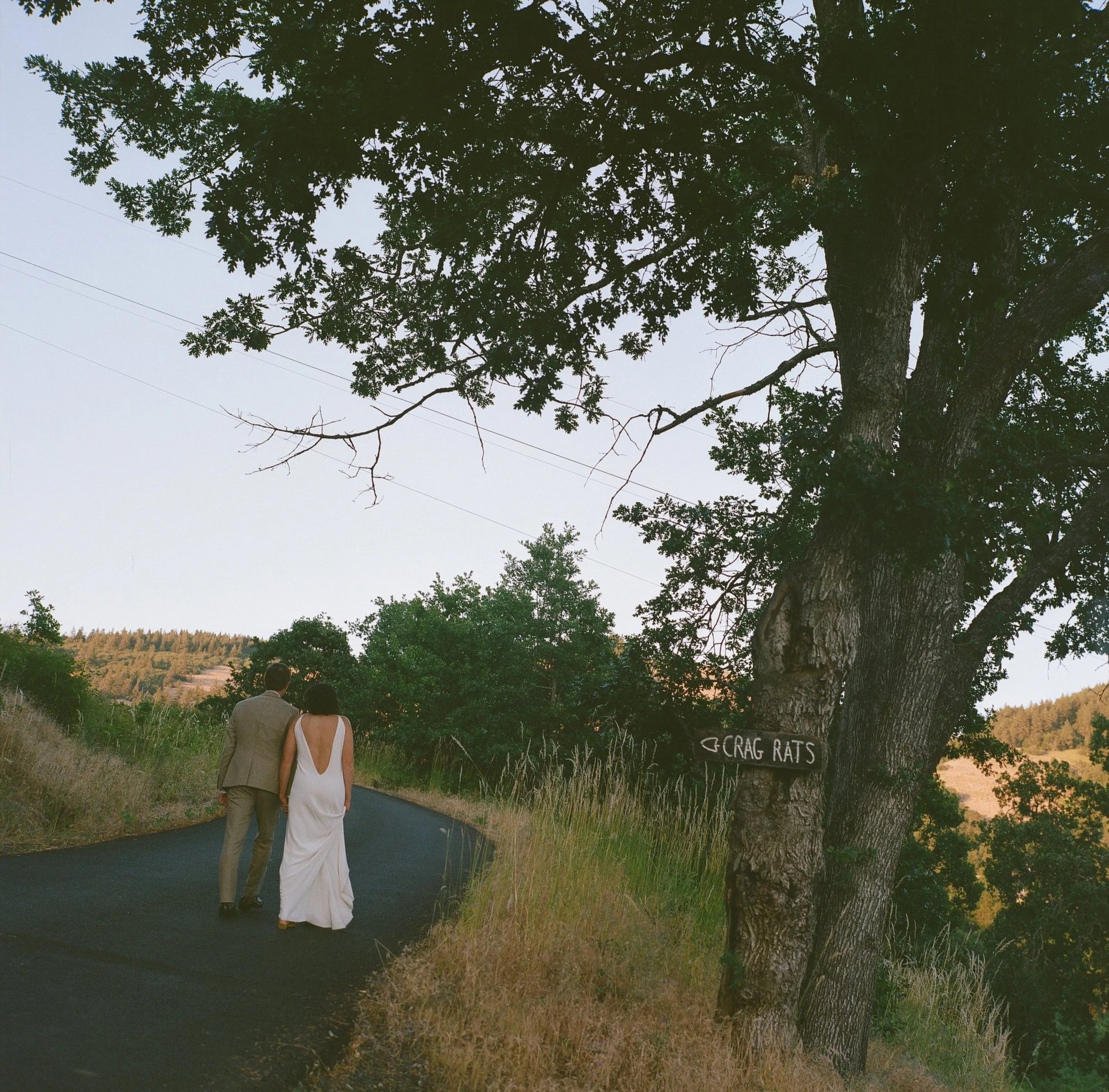 A couple dressed in formal attire walking on a rural road surrounded by grass and trees, with a sign on a tree pointing towards 'Crag Rats'.