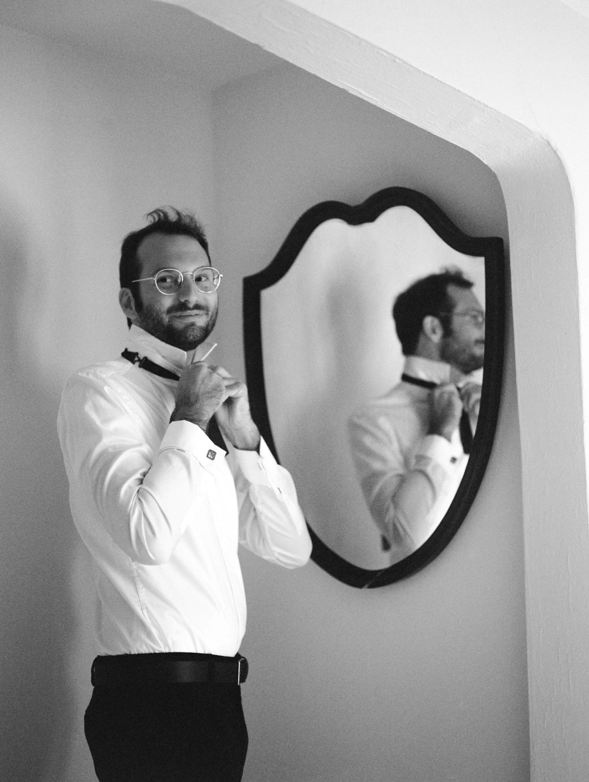 A man in a white dress shirt adjusting his bow tie while looking at a mirror on the wall.