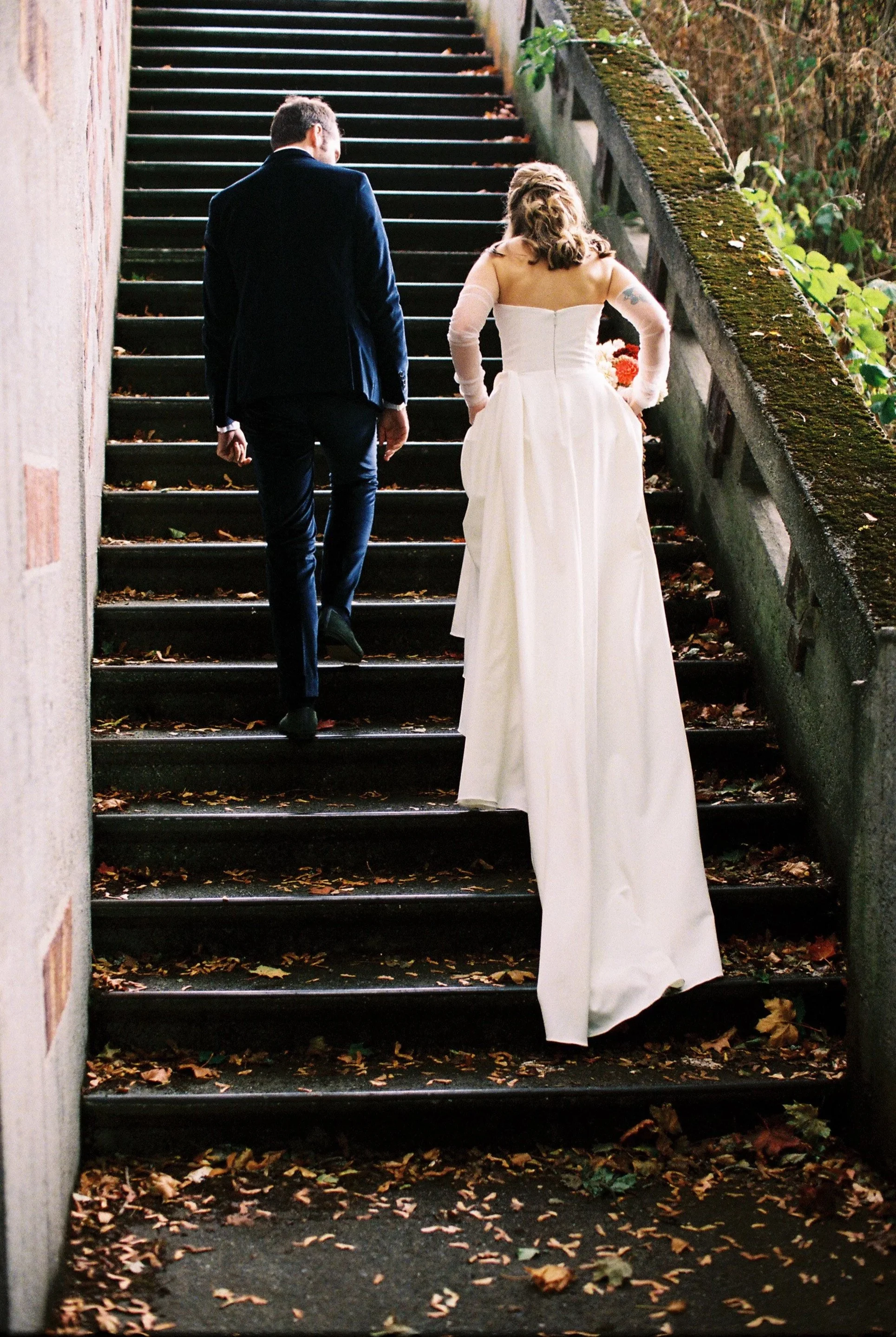 A couple, with the woman in a white wedding gown and the man in a dark suit, walking up outdoor stairs surrounded by autumn leaves.
