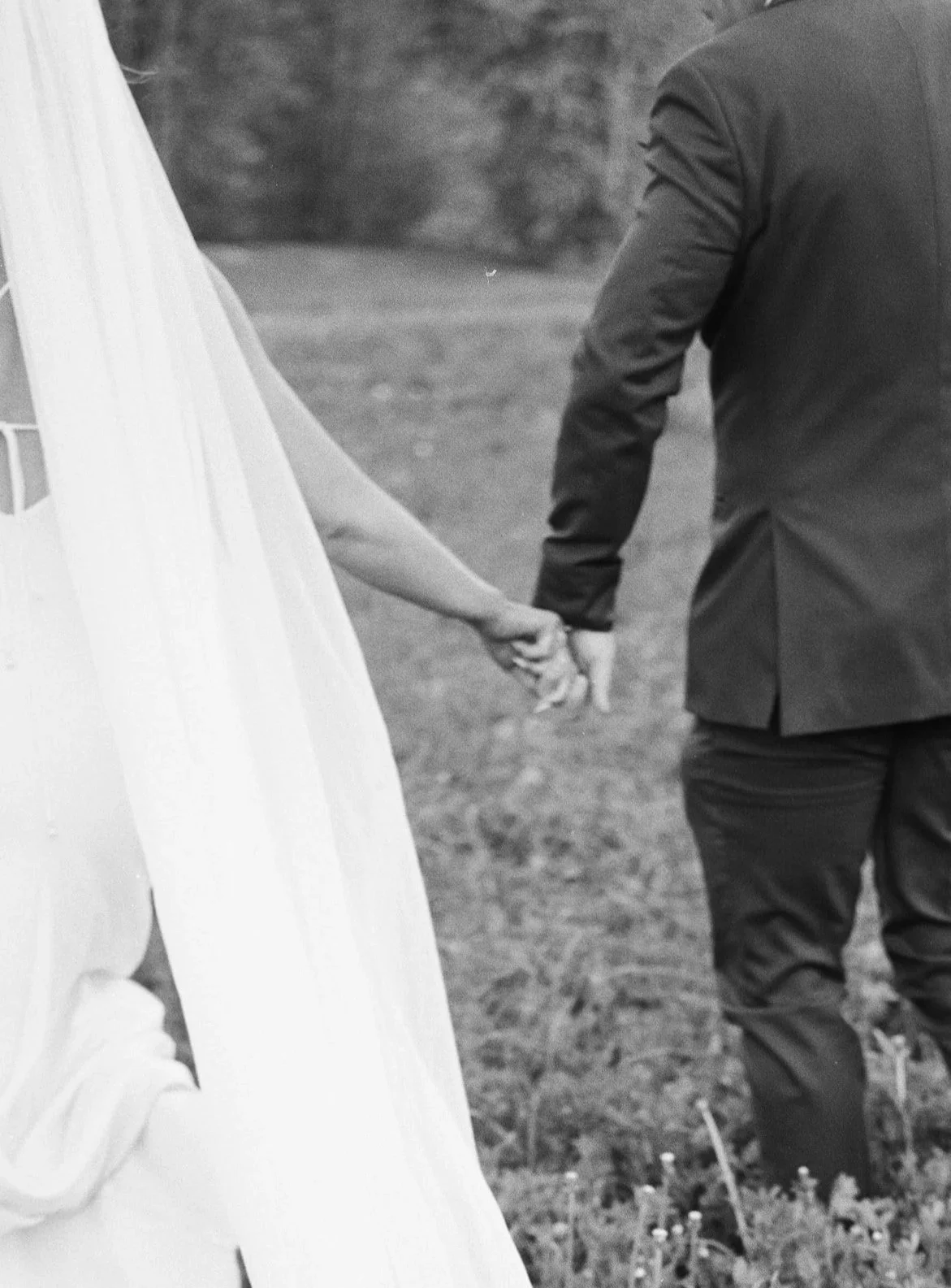 A black and white photograph of a bride and groom holding hands outdoors. The bride's dress is visible on the left, and the groom is wearing a suit. They are standing on grass with a blurred natural background.