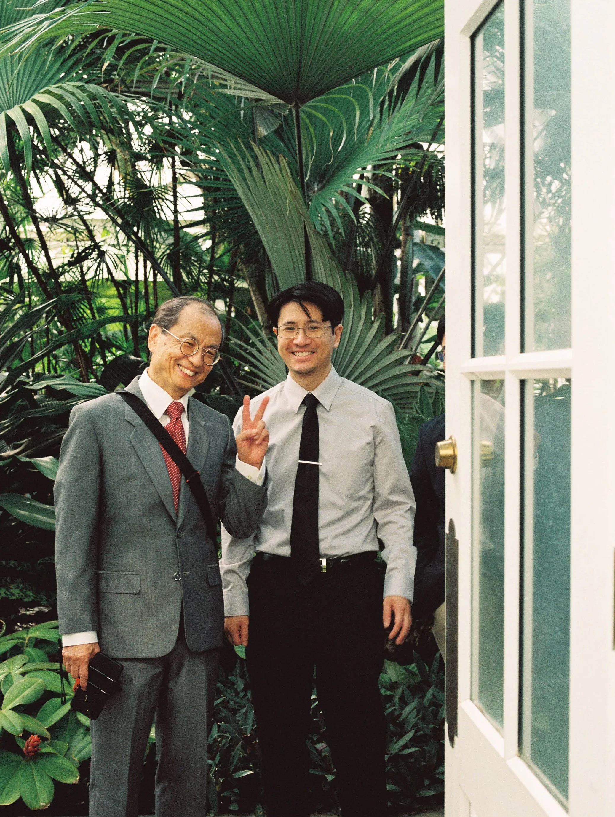Two men standing near a doorframe in front of large tropical green plants, smiling at the camera; one man is making a peace sign.