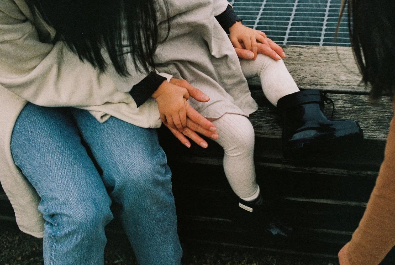 Two people sitting on a bench, one adult and one child, holding hands with their fingers intertwined. The child's legs are visible, wearing beige leggings and black shoes. The adult is wearing blue jeans and a light-colored jacket.