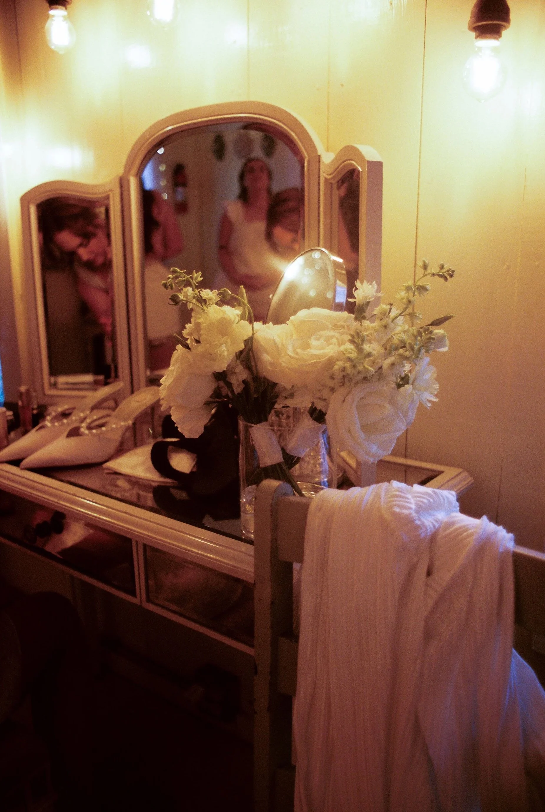 A vintage vanity with a bouquet of white roses and flowers, a mirror, and a pair of white shoes on the surface, with a yellow wall and light bulbs overhead, and a blurred reflection of three women in the mirror.