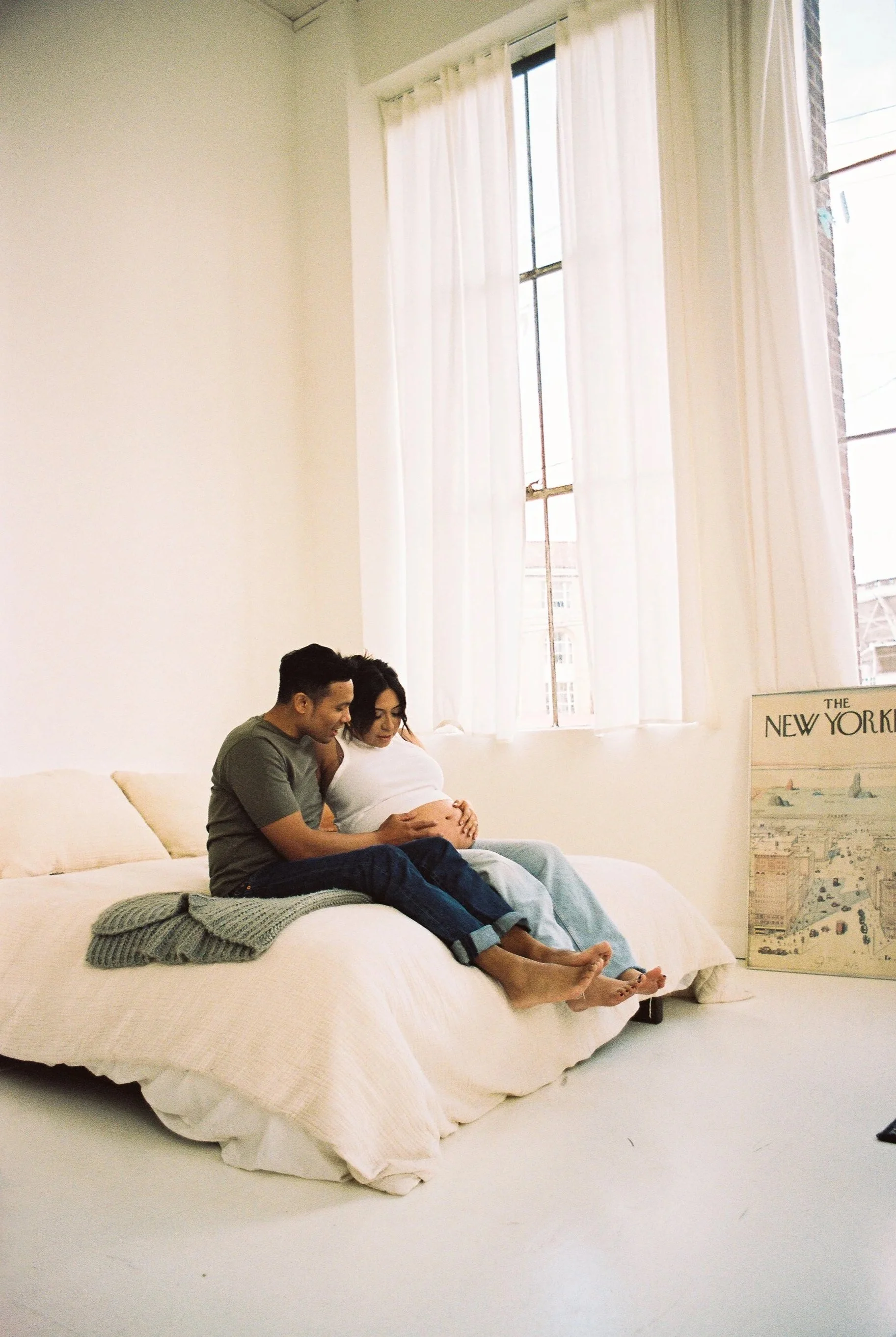 A couple sitting on a bed, with the woman pregnant, looking at her stomach.