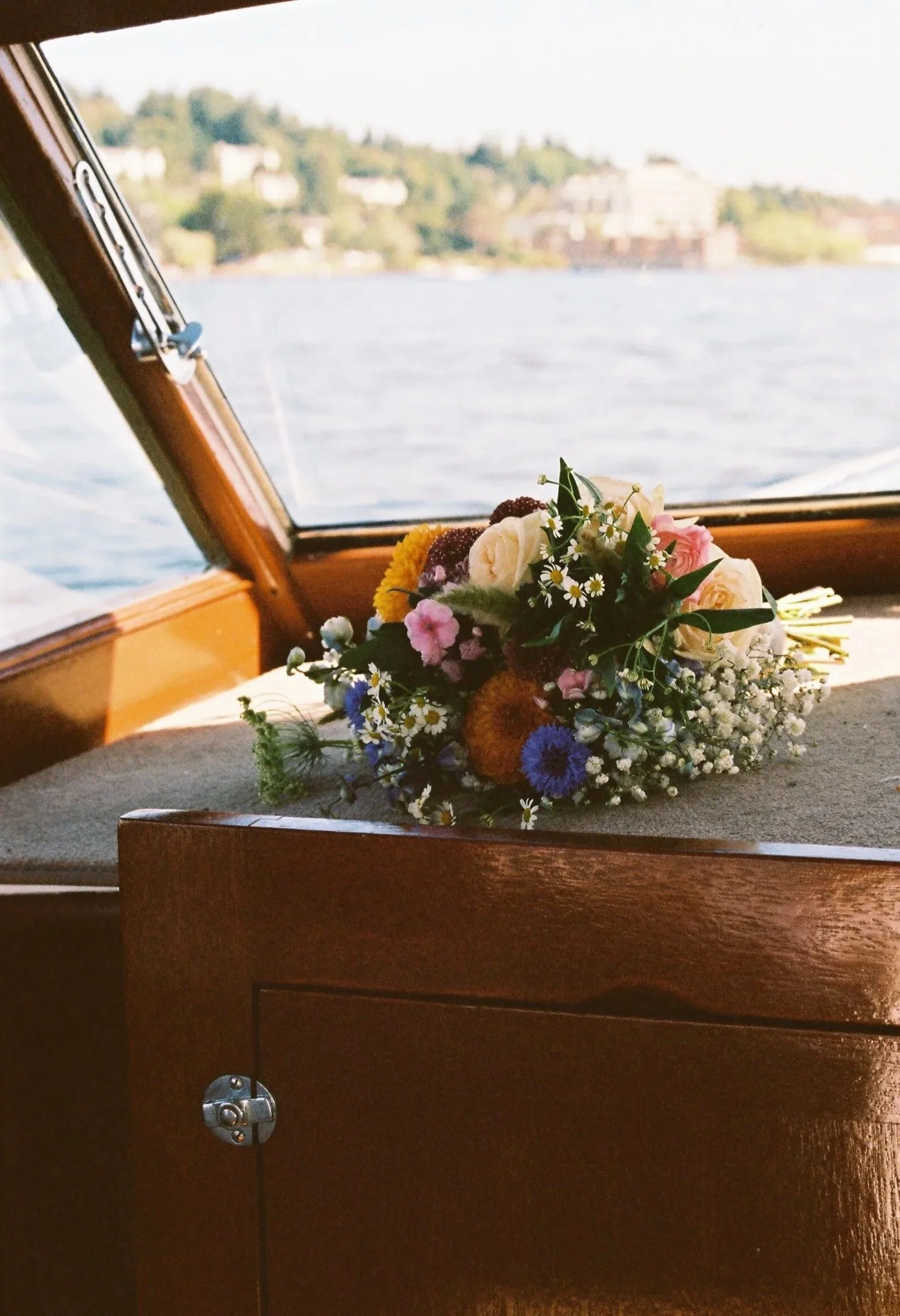 A bouquet of multicolored flowers resting on a wooden surface inside a boat with water and a shoreline with buildings in the background.