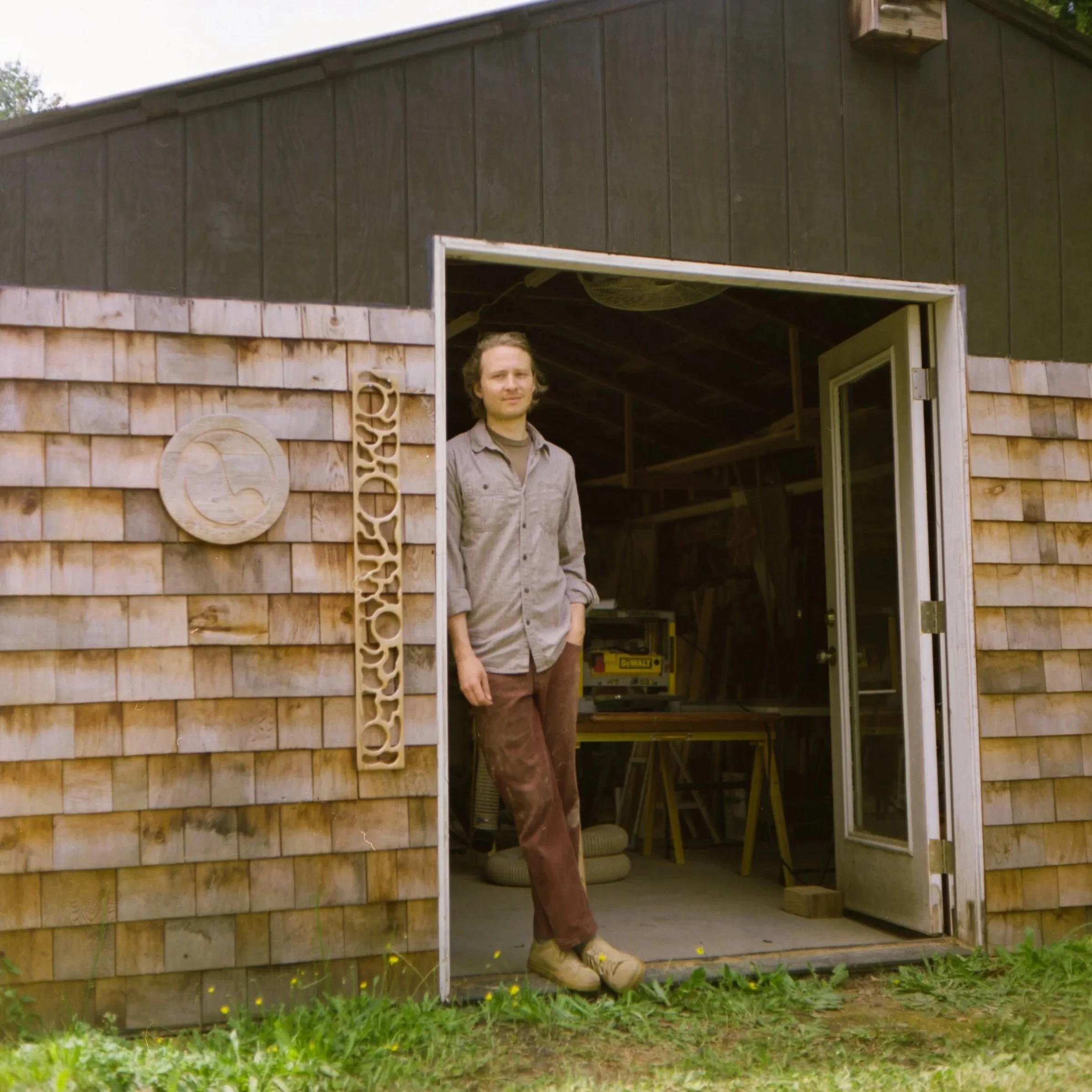 A man stands in the doorway of a wooden shed, leaning slightly with his hands in his pockets, inside the shed there are tools and equipment, with a sign that says 'BYO' outside the shed.