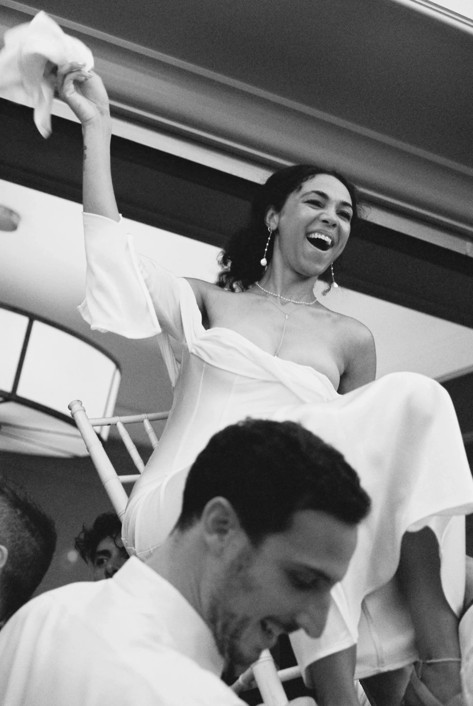 A woman in a white dress, sitting on a chair, laughing and holding a cloth, surrounded by people at a celebration or event.