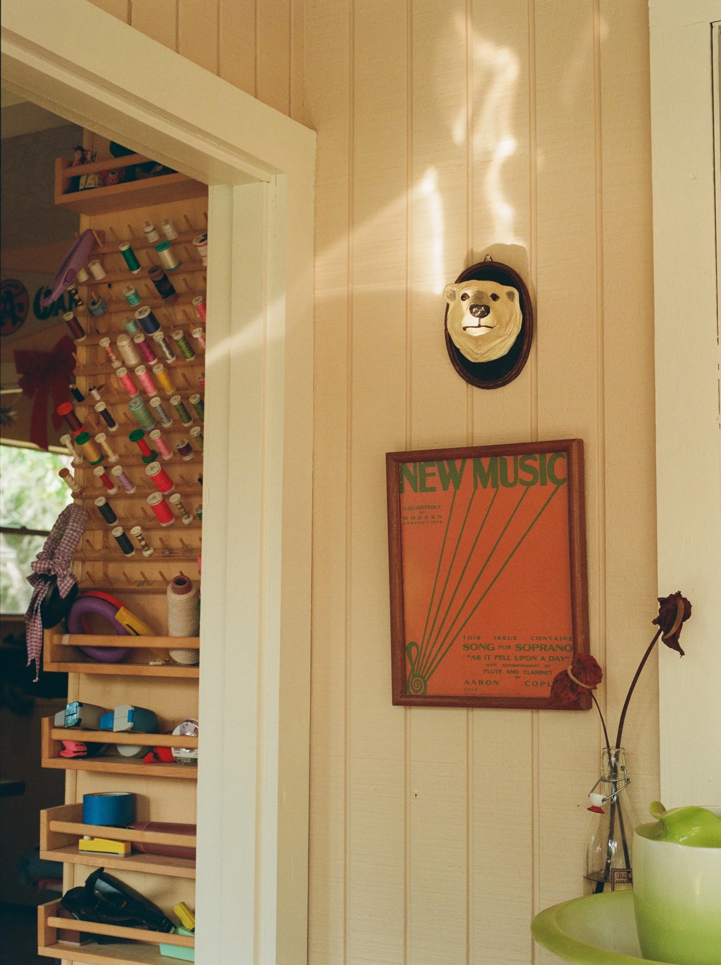 A wall with a mounted bear mask, a framed vintage music sheet, and a vase with dried roses, with a doorway to a craft area on the left showing spools of thread and tape.