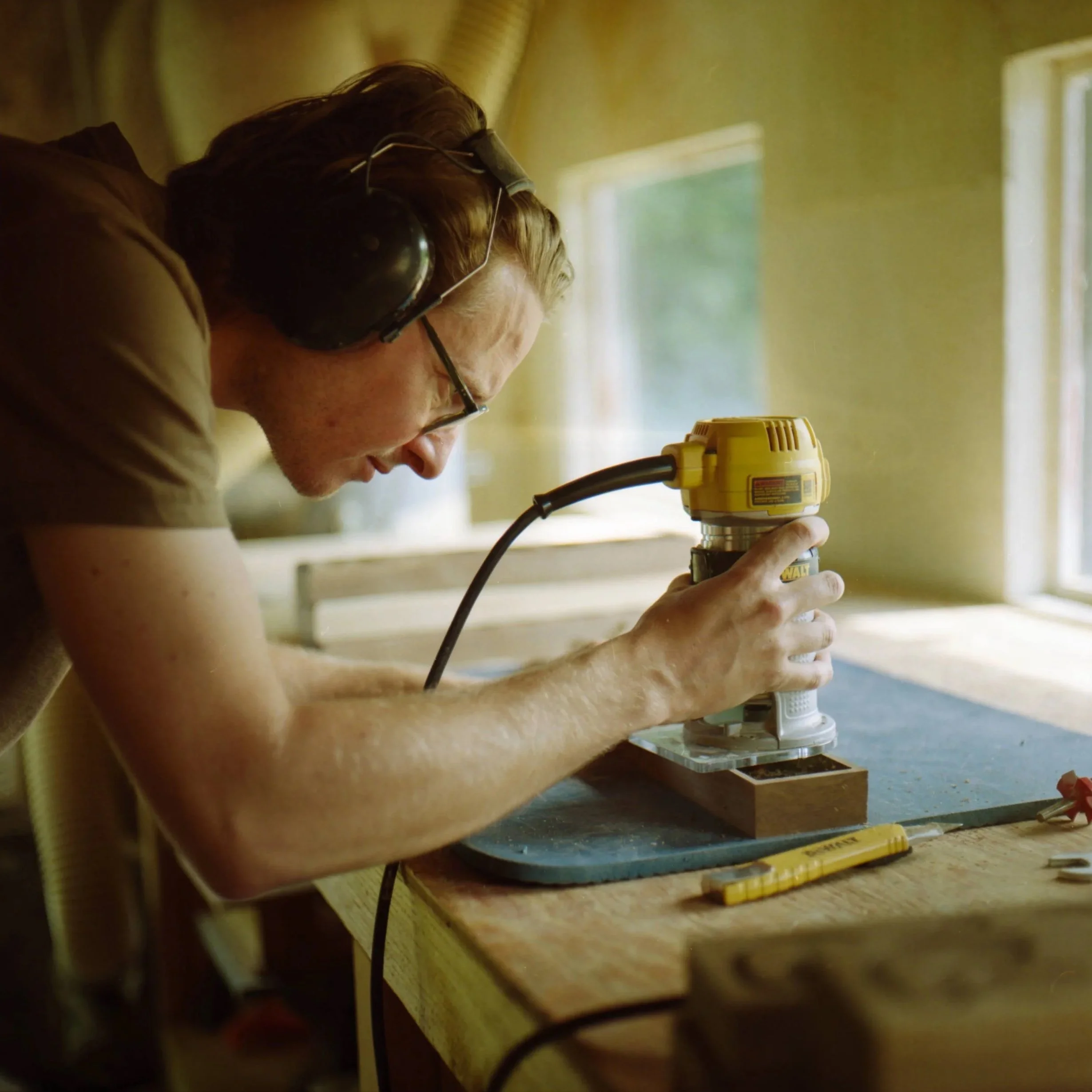 Man wearing glasses and earmuffs focused on using a handheld power tool on a piece of wood in a workshop with sunlight coming through the window.