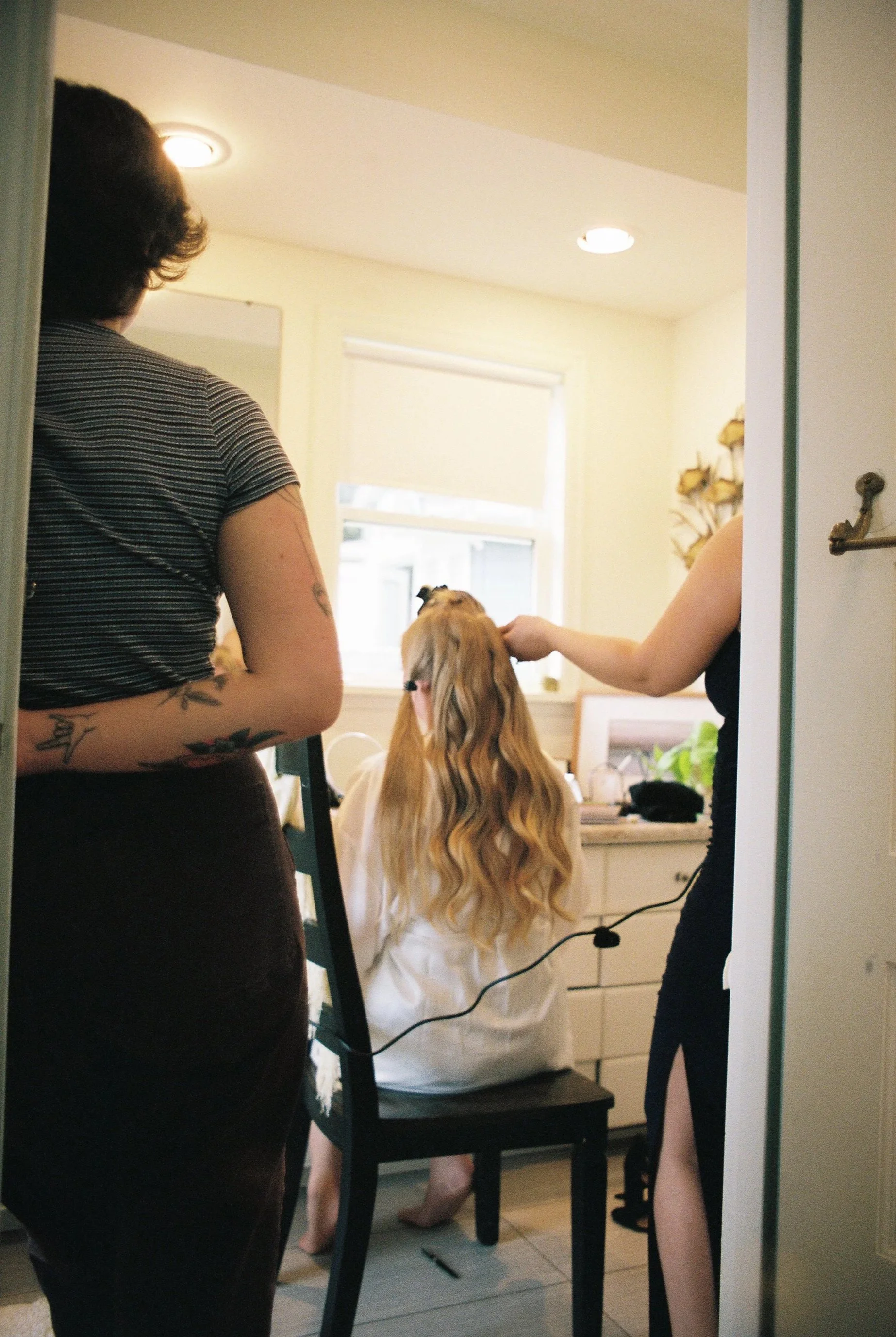 A woman with long blonde hair getting her hair styled while sitting on a chair in a well-lit room, with another woman and a person with short curly hair attending to her.