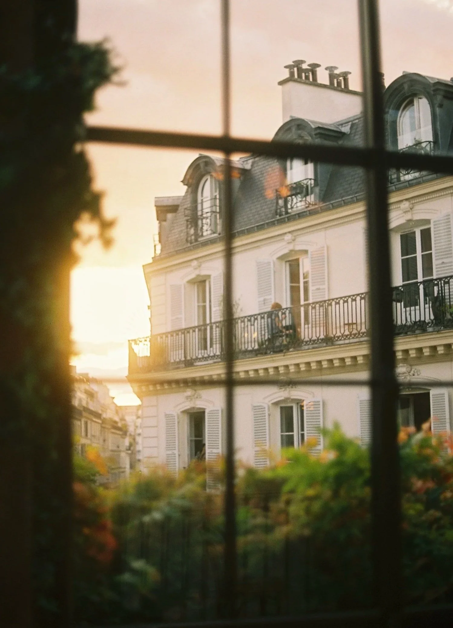 View of a classic Parisian apartment building with white shutters and wrought-iron balconies, seen through a window with bars, during sunset.