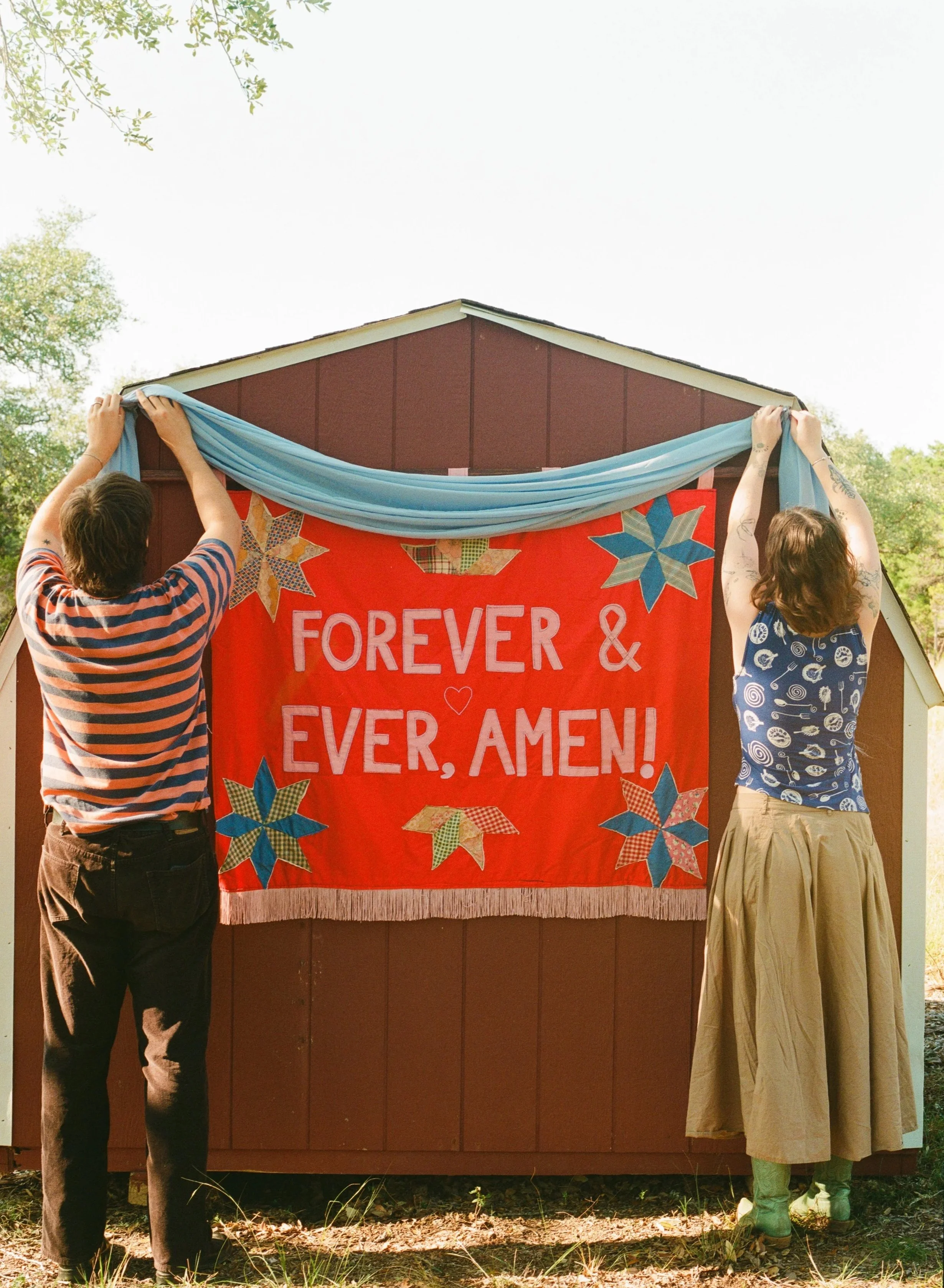 Two people hang a textile banner with the words "Forever & Ever, Amen!" on a small outdoor building, with a blue fabric draped on top. The banner is red with multicolored star patterns, and the scene is outdoors with trees in the background.