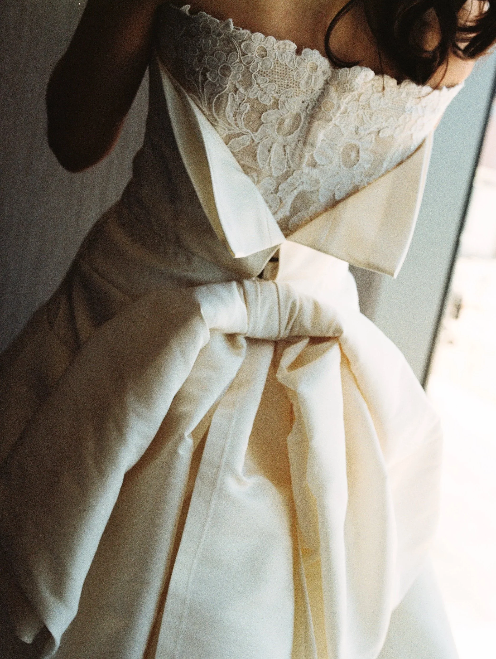Close-up of a person in a cream-colored dress with lace embroidery, sitting or leaning near a doorway.