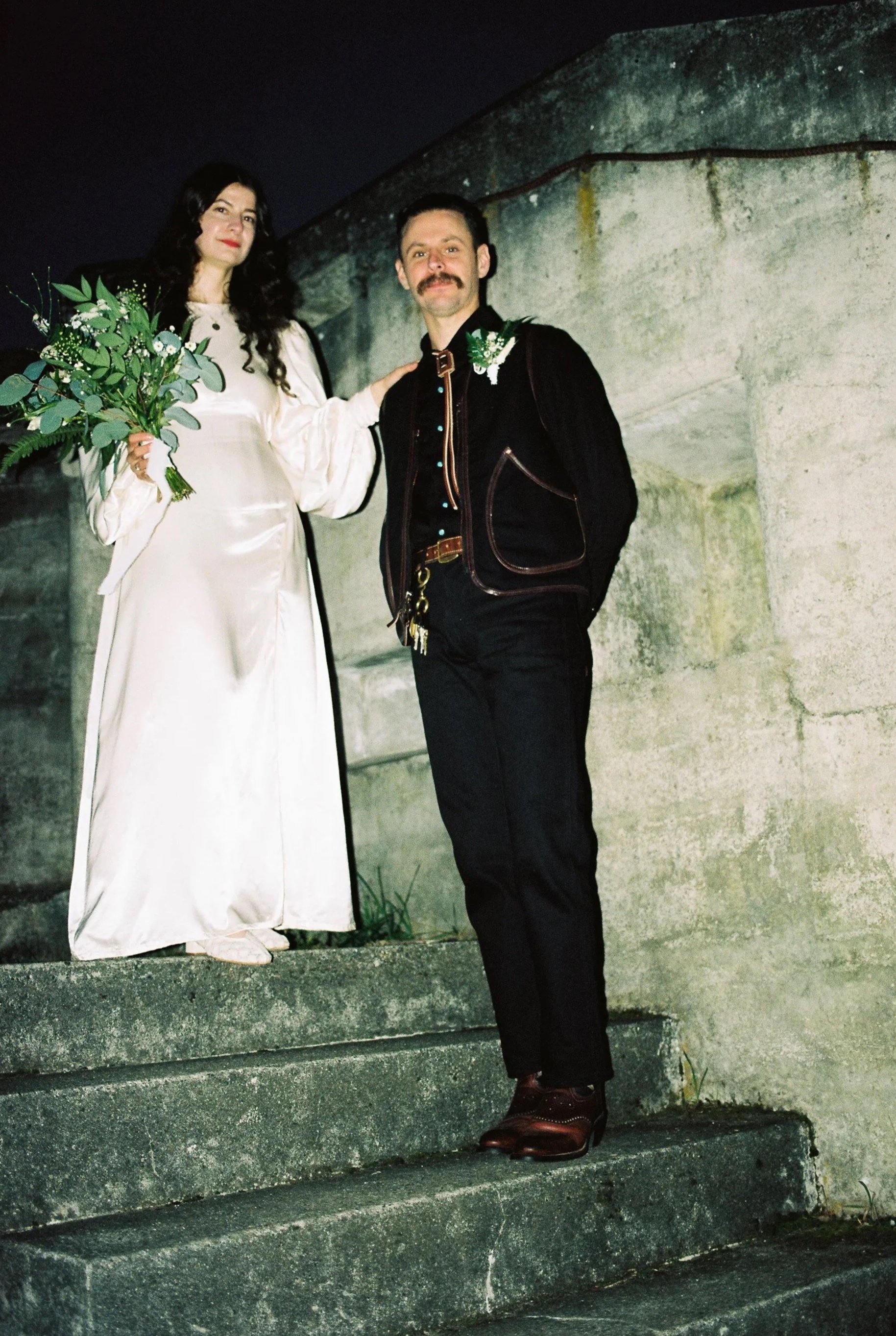 A couple stands on outdoor stone stairs at night, the woman in a white dress holding a bouquet, and the man in dark clothing with a mustache.