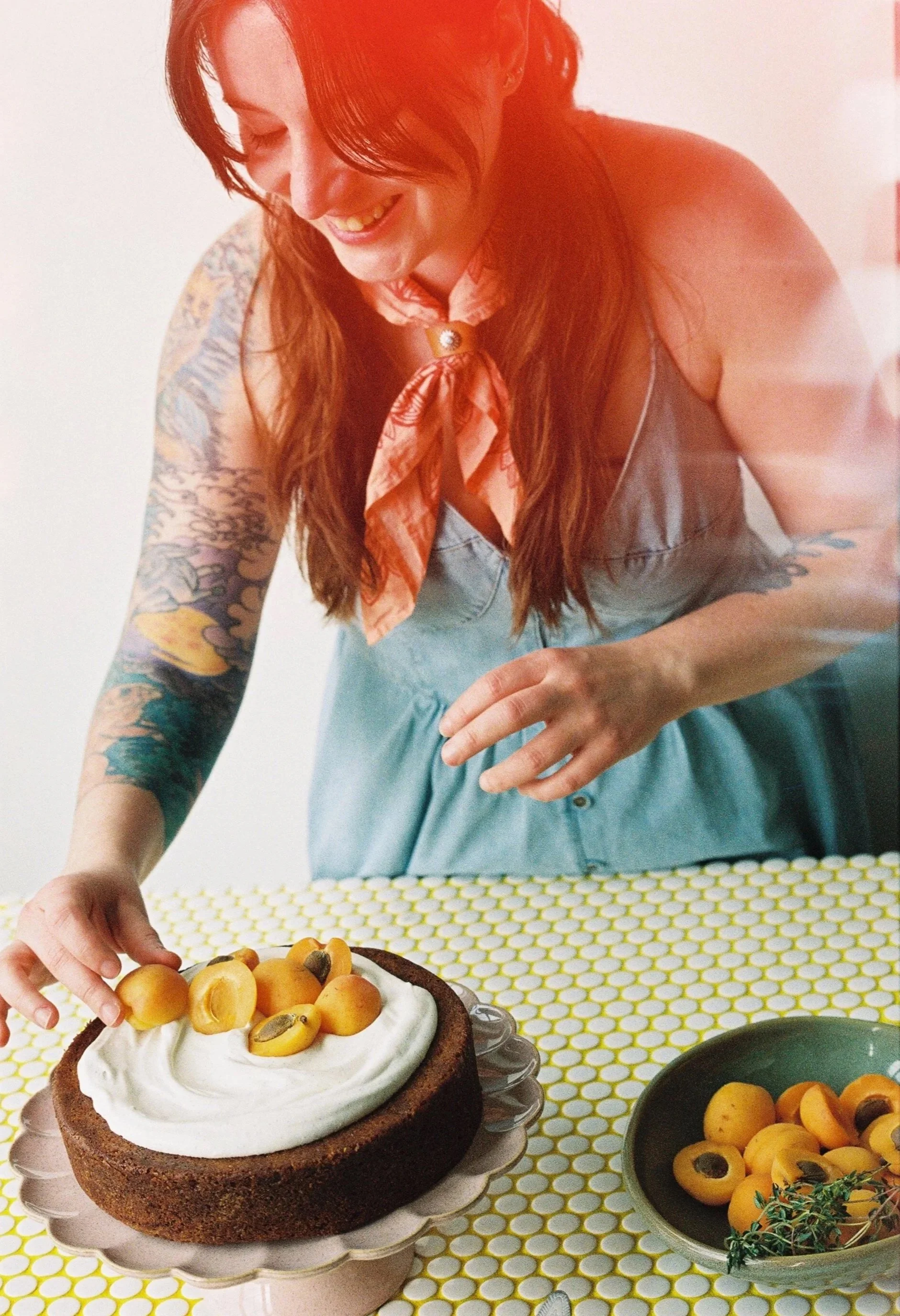 Woman with long red hair, tattoos on her arms, and wearing a denim dress and pink scarf, decorating a cake with apricots and whipped cream on a yellow polka dot tablecloth.