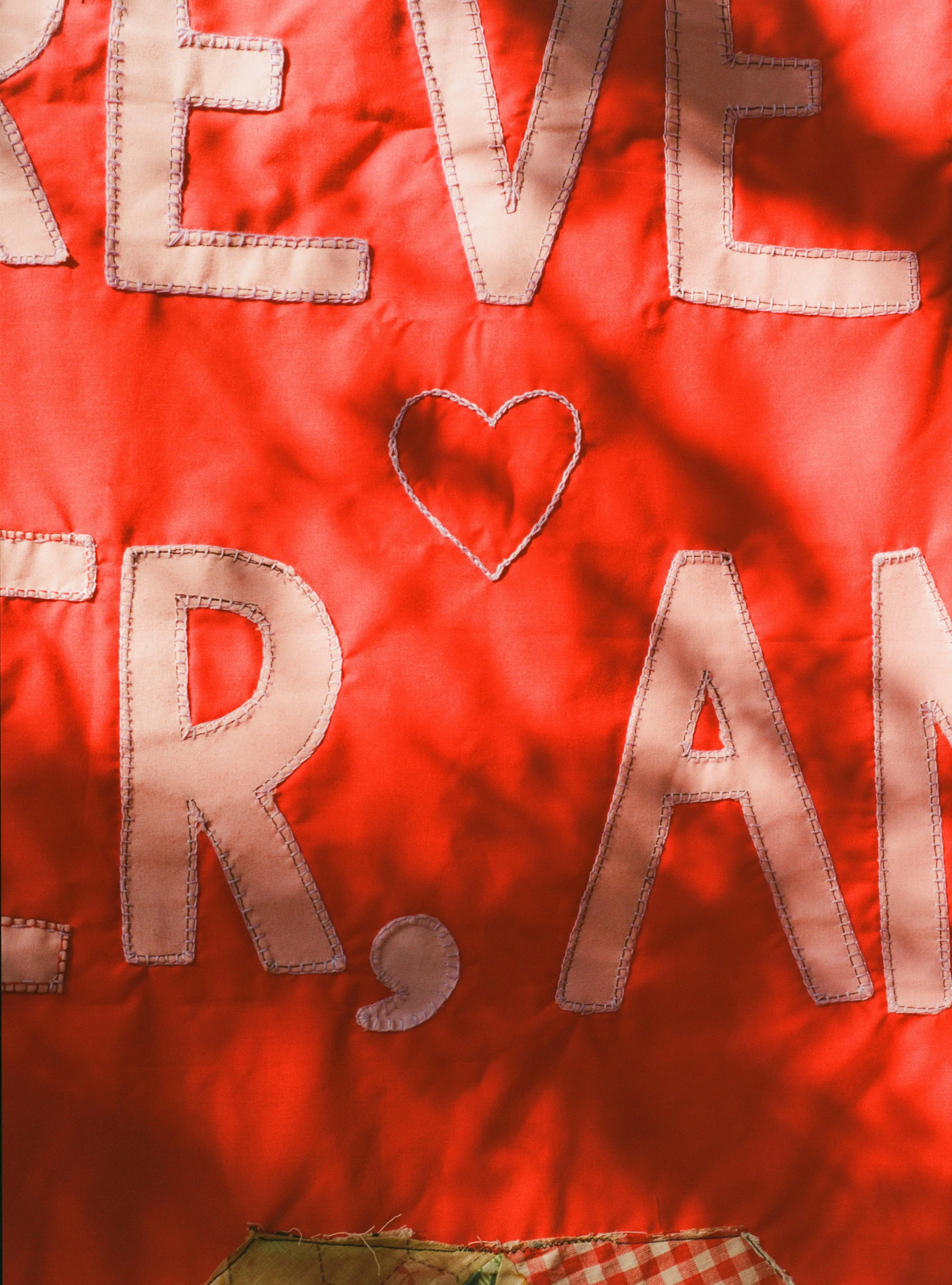 Red fabric background with beige stitched letters spelling 'LOVE' and 'READ' arranged around a stitched heart shape.